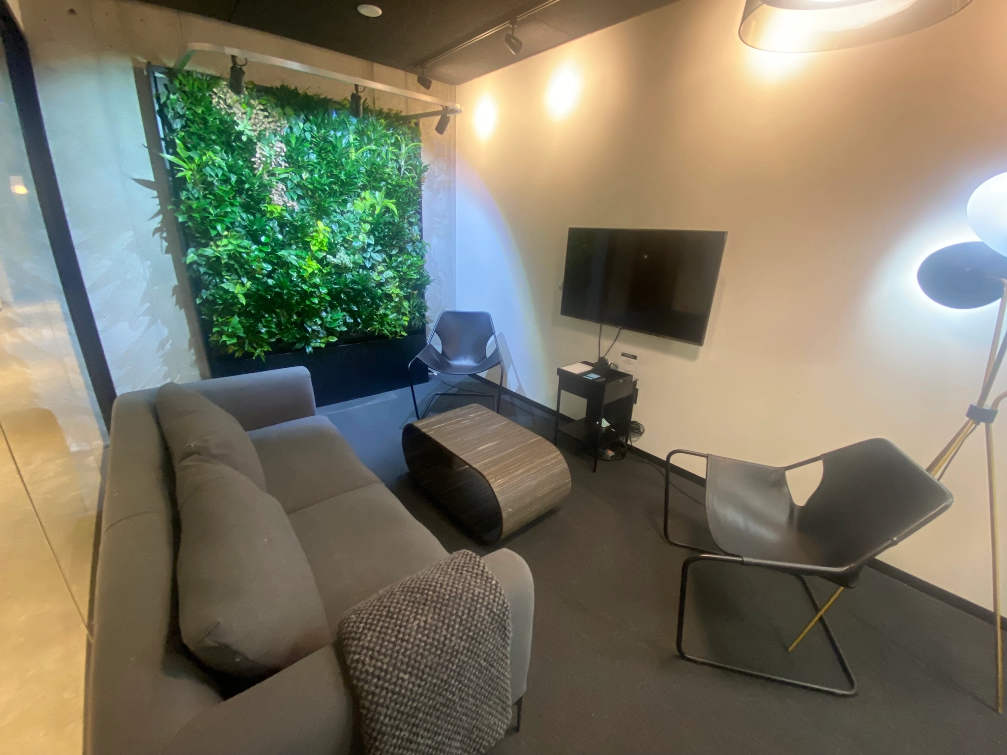 A modern coworking lounge area with a gray sofa, two chairs, a coffee table, a wall-mounted TV, a floor lamp, and a wall adorned with green plants.