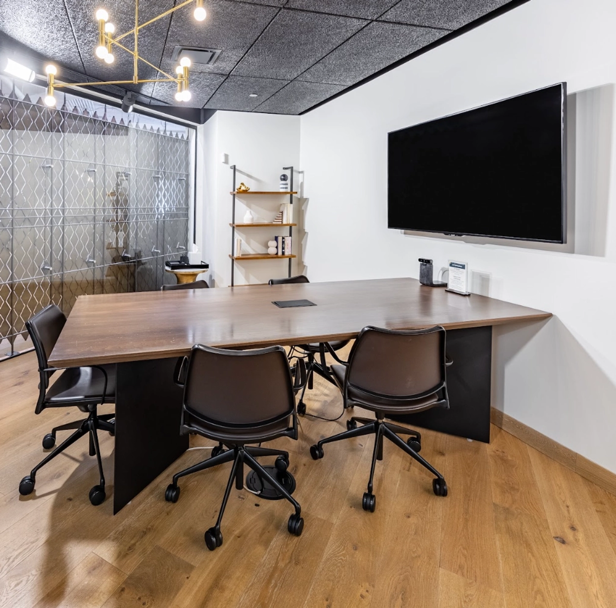 A modern coworking space with a large wooden table, five black office chairs, a wall-mounted flat-screen TV, wooden shelving, and a geometric pendant light fixture.