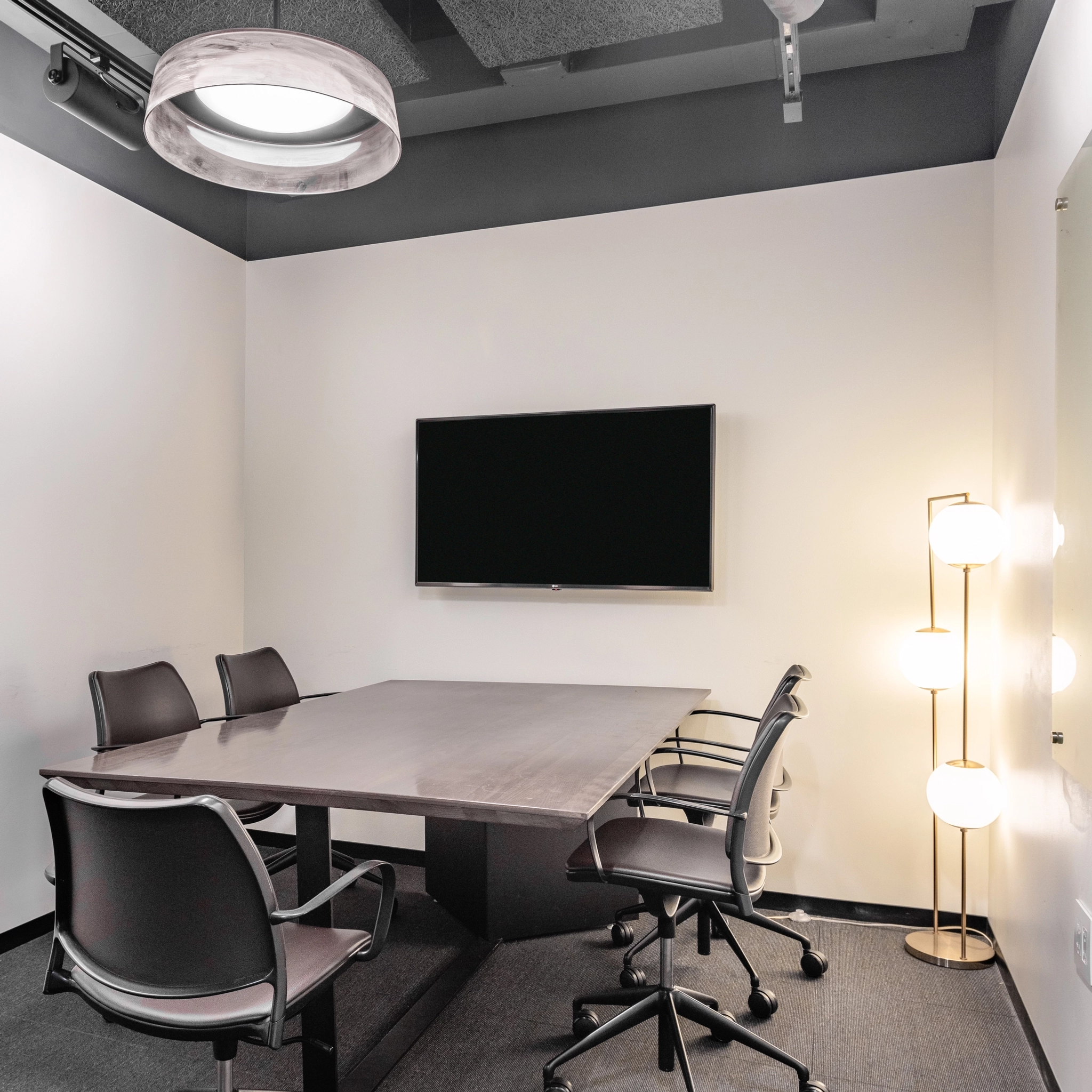 A small conference room in the McLean office features a table, four chairs, a wall-mounted TV, and a floor lamp with three globe lights.