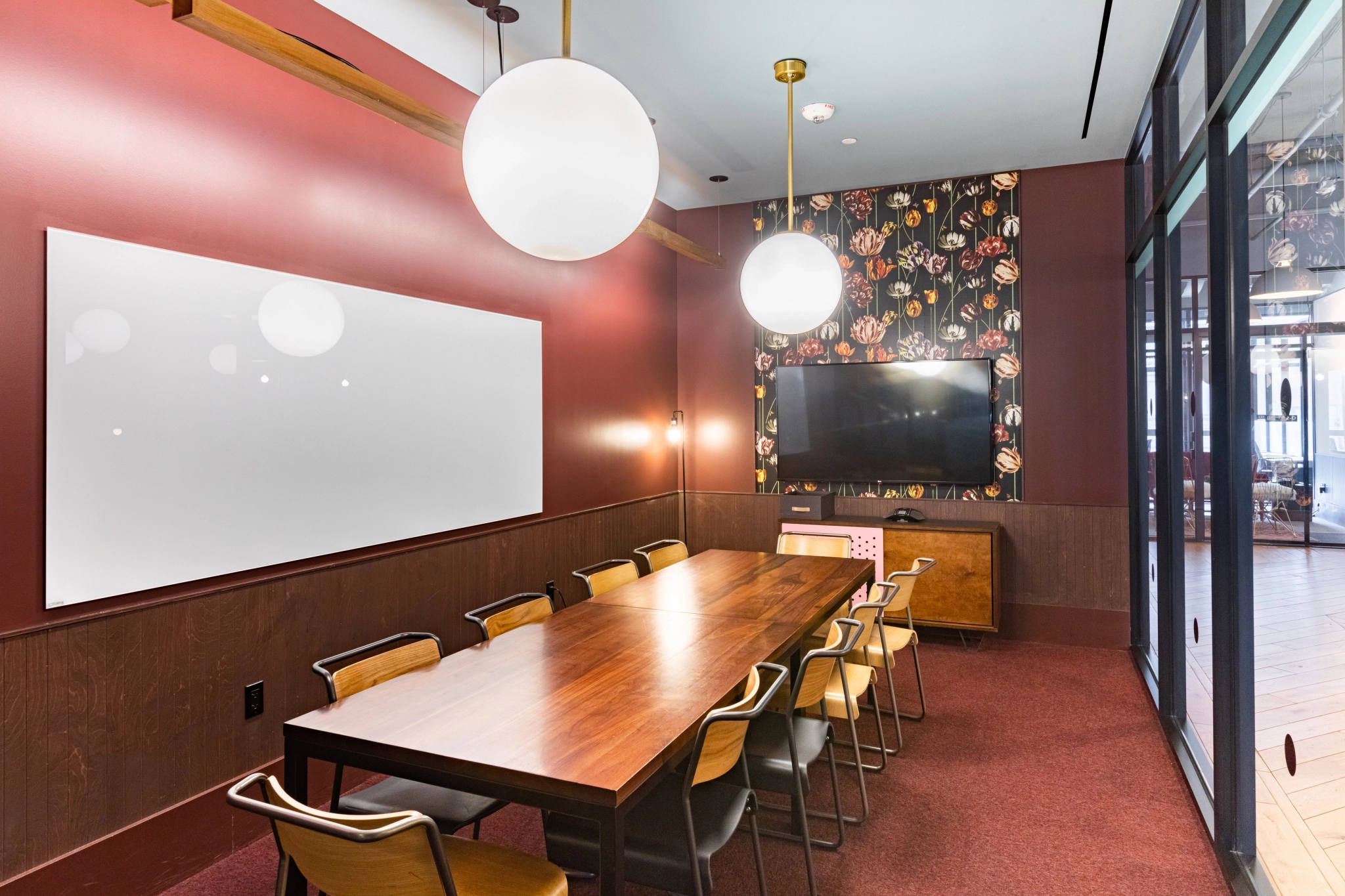 A modern meeting room with a long wooden table, chairs, a large whiteboard, flat-screen TV, and spherical ceiling lights. This office in Washington features burgundy walls and a large window on the right side.