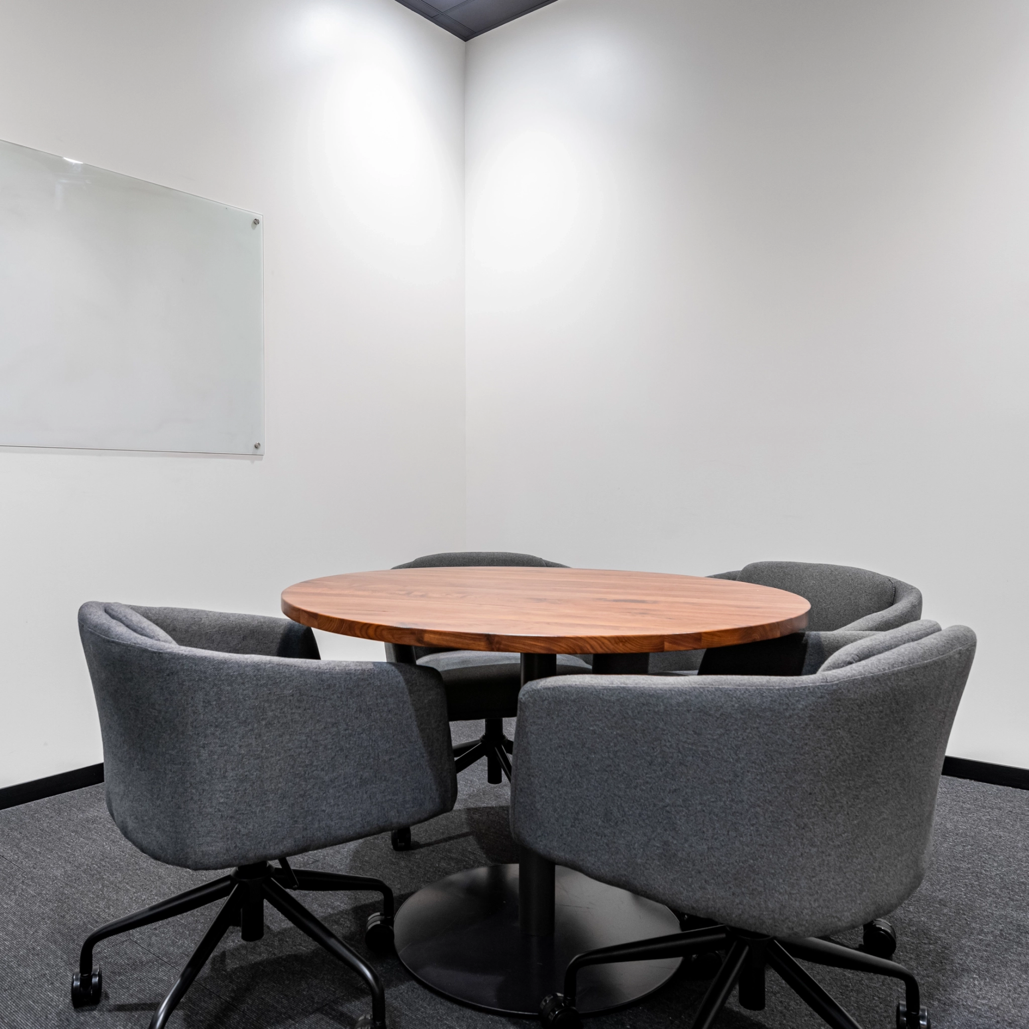 A small meeting room in Walnut Creek features a wooden round table, four gray chairs, and an empty whiteboard set against a crisp white wall, making it an ideal office space for collaboration.