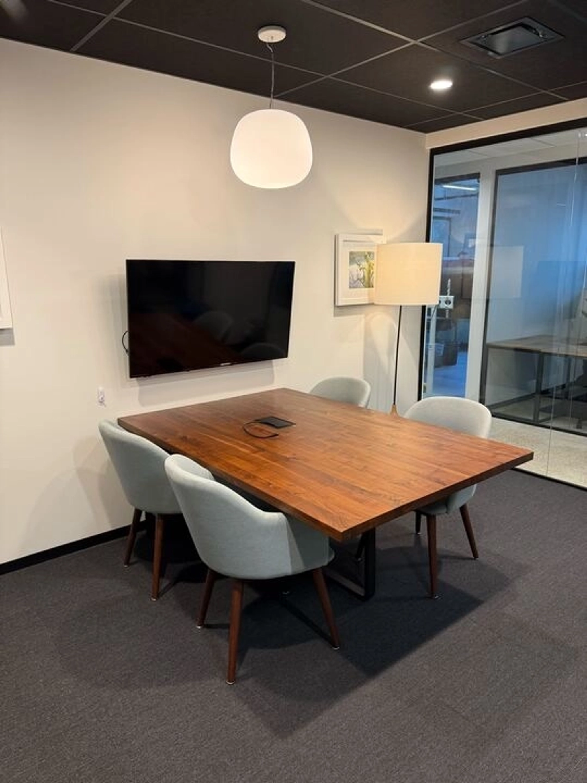 Ein kleines Besprechungszimmer in Washington mit einem Holztisch, vier grauen Stühlen, einem Wandfernseher, einer Stehlampe und gerahmten Kunstwerken an der Wand.