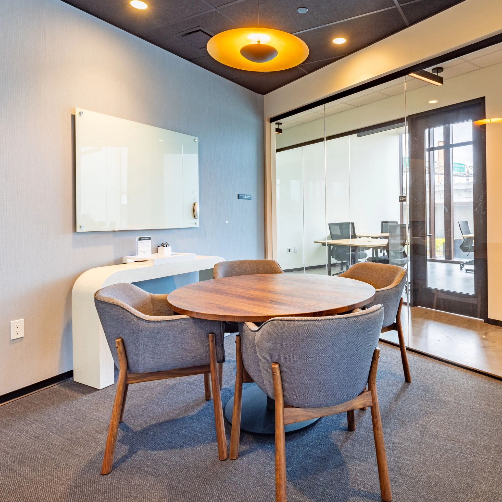 A small meeting room with a round wooden table, four gray chairs, a whiteboard, and a large window looking into another coworking area.