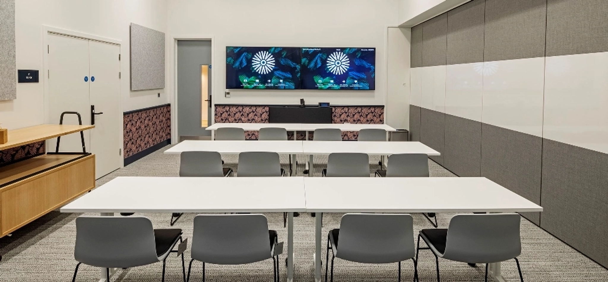 A modern classroom or meeting room in Manchester with three white tables, gray chairs, wall-mounted screens at the front, and neutral decor.