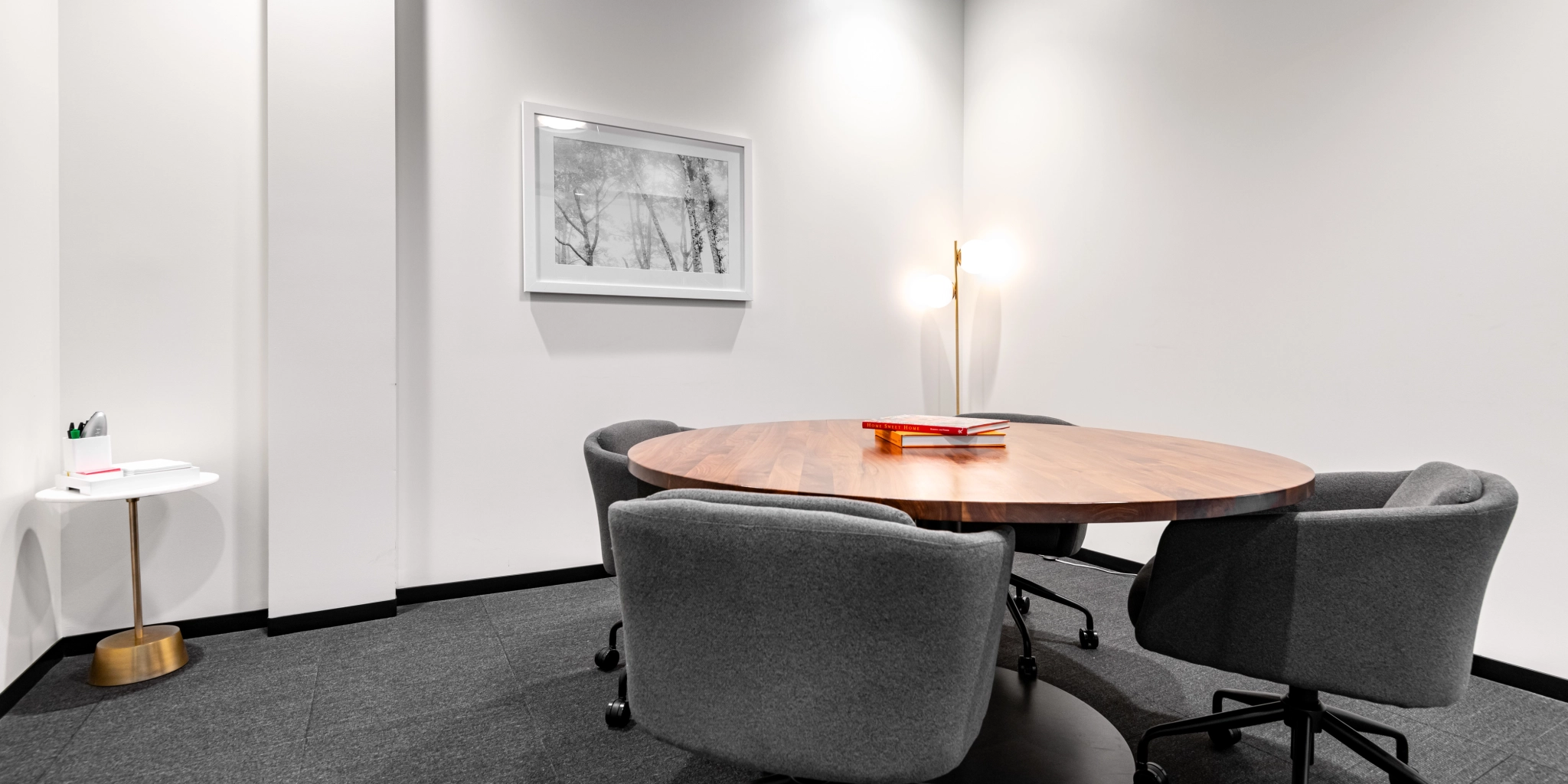 A round wooden table with four gray chairs anchors the cozy workspace in a Walnut Creek meeting room. A small side table with pens rests on the left, while a framed picture adorns the crisp white wall, creating an inviting atmosphere for coworking.