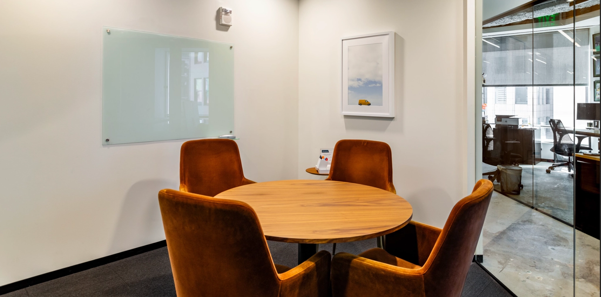 Une petite salle de réunion avec une table ronde en bois, quatre chaises marron rembourrées, un tableau blanc au mur et une photo encadrée, idéale pour les séances de coworking. Cet espace de travail confortable est adjacent à un espace de bureau ouvert plus grand dans la ville animée de San Francisco.