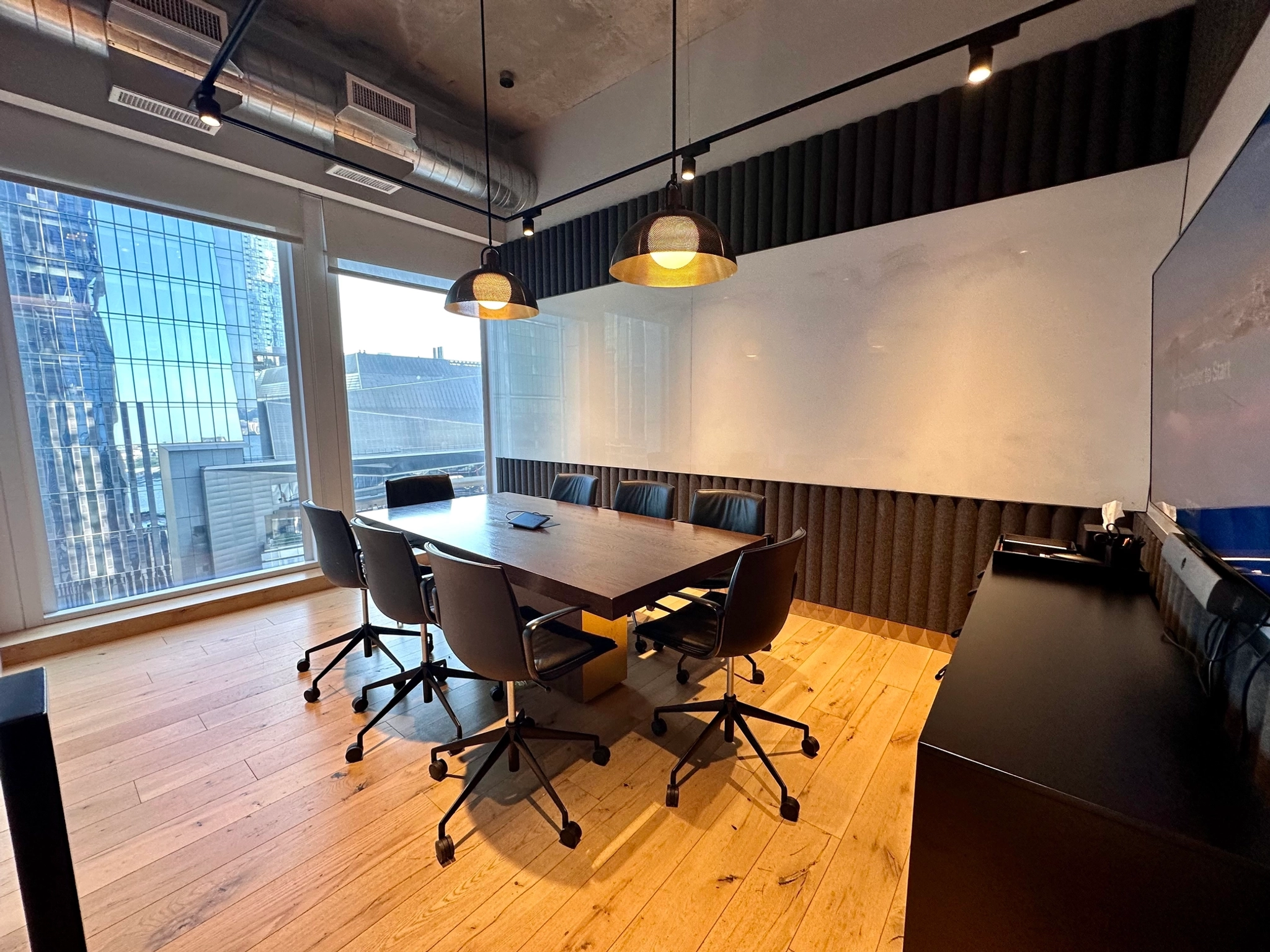 A modern meeting room with a wooden table, six chairs, a whiteboard, and pendant lights overlooks New York's skyscrapers through large windows.