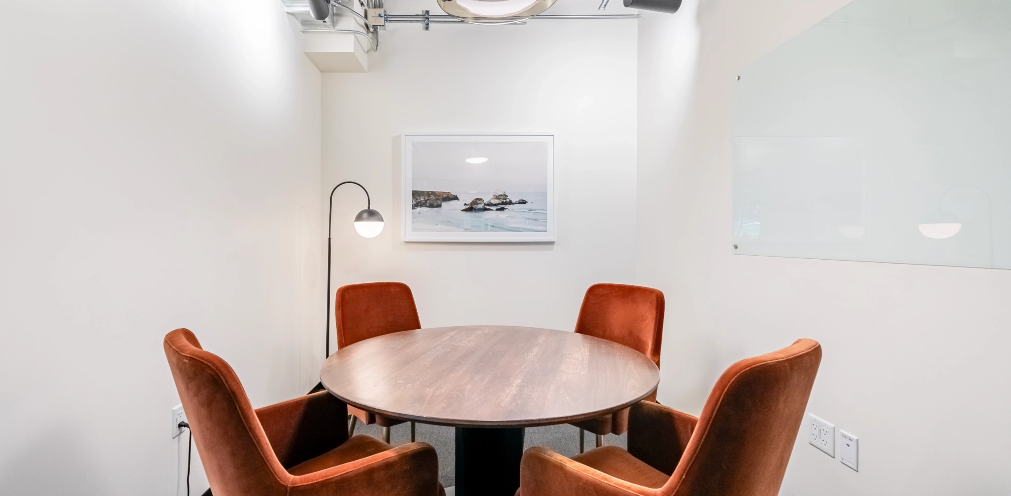 Une petite salle de réunion dans un bureau de San Francisco comprend une table ronde en bois, trois chaises de couleur rouille, un lampadaire et une photographie murale d'une scène de plage.