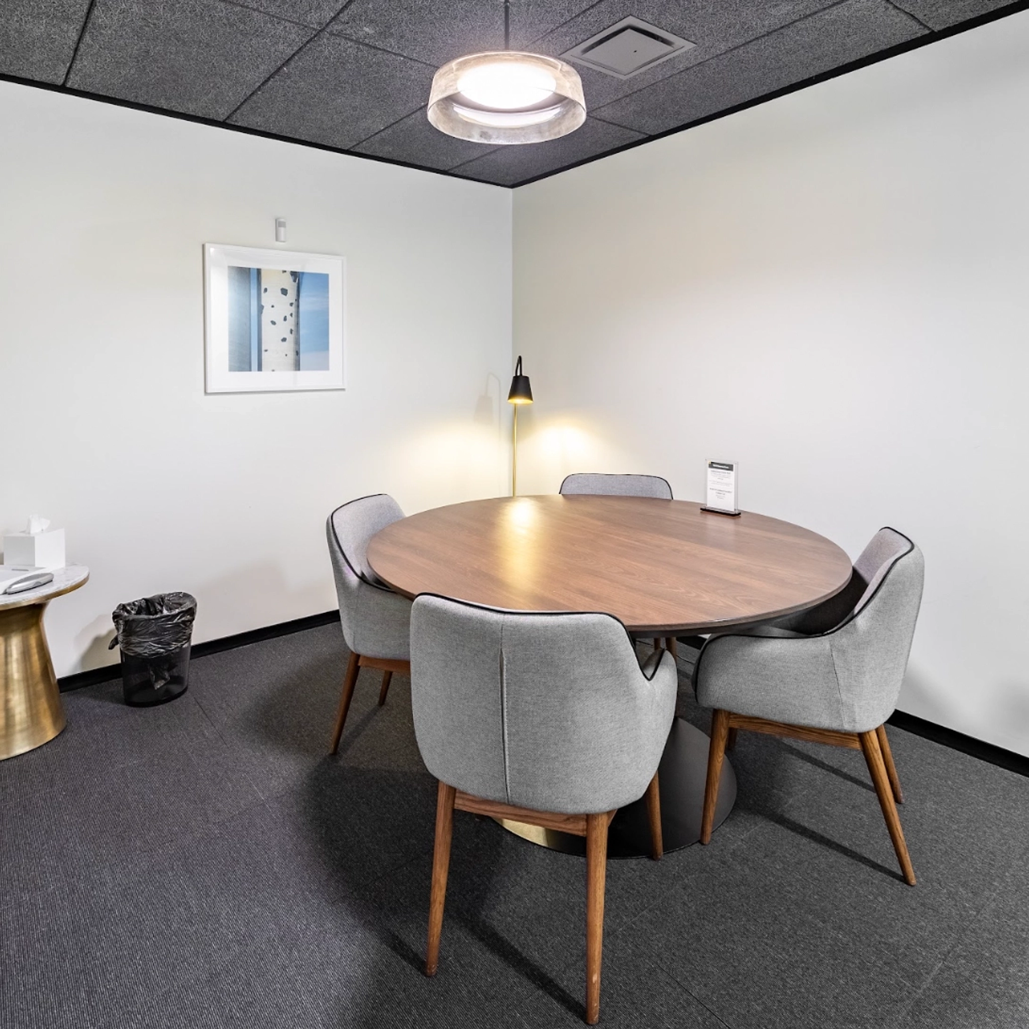 A small coworking meeting room with a round table, four gray chairs, a hanging light fixture, wall-mounted artwork, and a side table with a tissue box and trash can. This workspace is perfect for collaborative discussions.