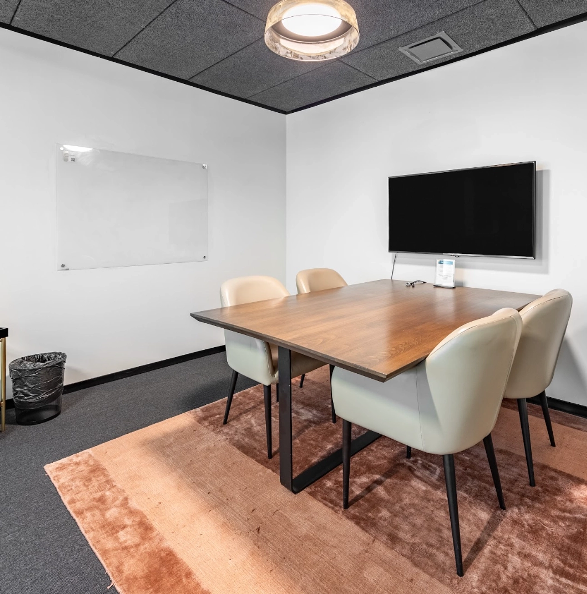 A small meeting room in the Arlington office features a wooden table, four beige chairs, a wall-mounted TV, a whiteboard, and a trash bin. The workspace is carpeted with a light brown rug.