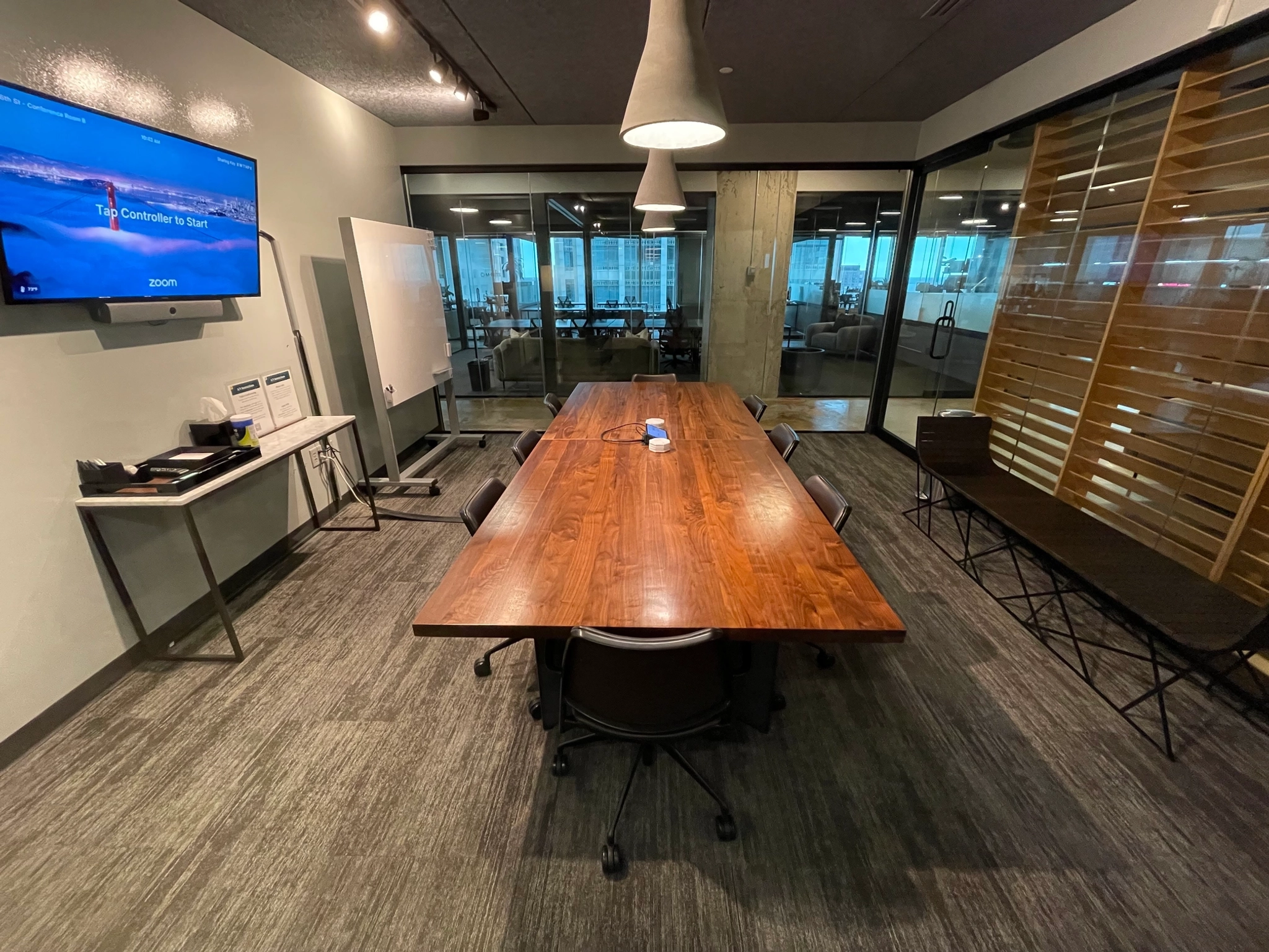 A modern meeting room with a long wooden table, black chairs, a tv display screen, and glass walls.