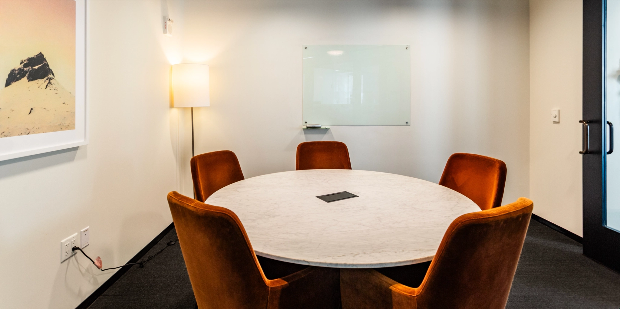 A small, elegant meeting room in San Francisco features a round marble table, four brown chairs, a whiteboard on the wall, a floor lamp, and a framed picture. Ideal for coworking sessions or intimate discussions.