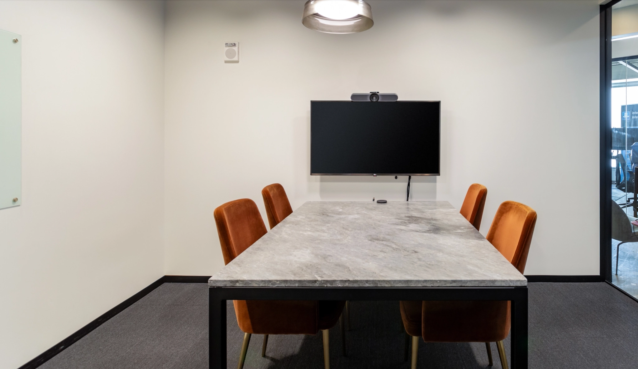 A small office meeting room with a rectangular marble table, four orange chairs, a wall-mounted TV, and a ceiling light. The workspace has white walls and a glass panel on one side.