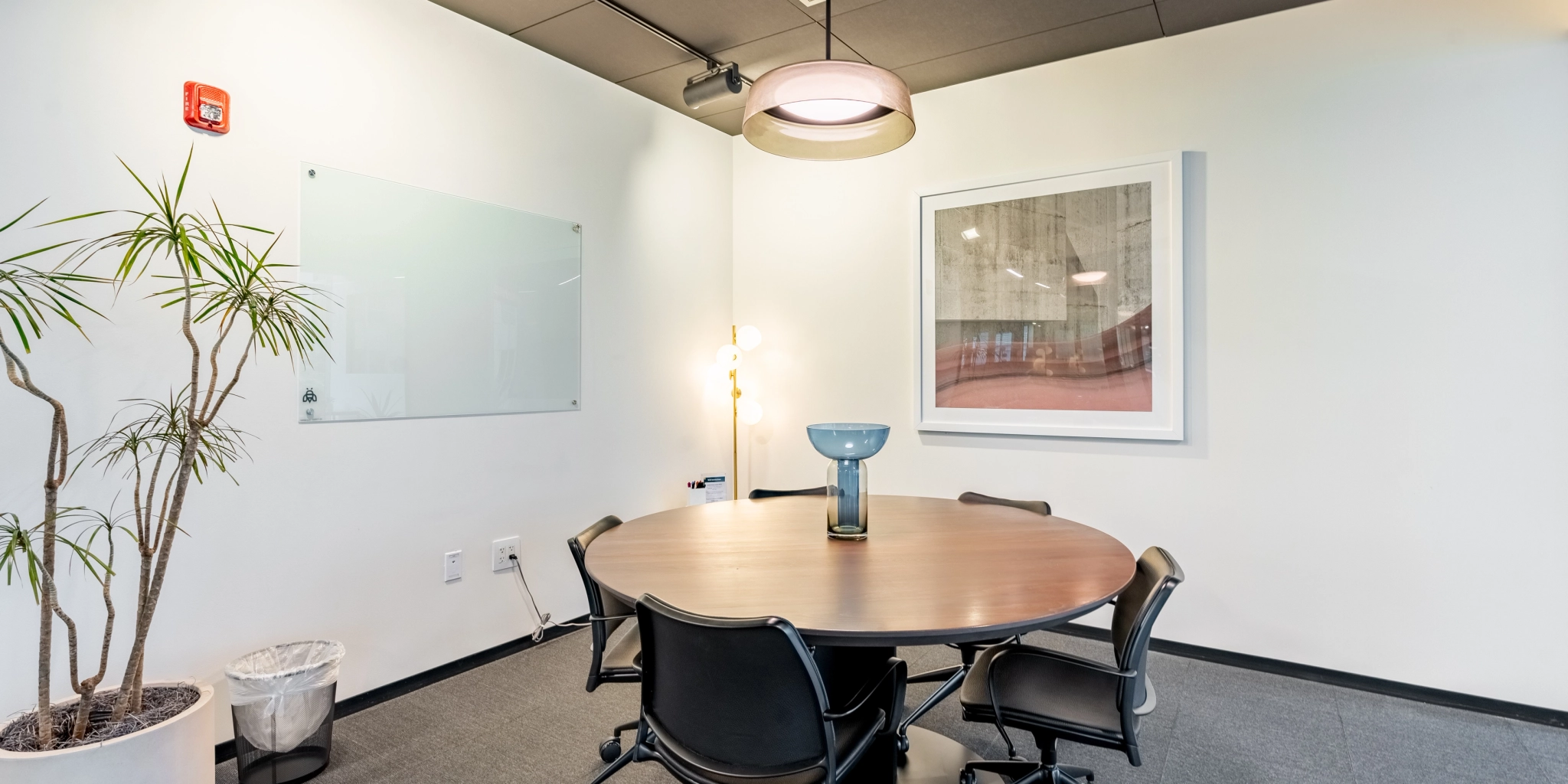 Small Arlington office conference room with a round table, four chairs, a whiteboard, a potted plant in the corner, and a framed picture on the wall. A pendant light hangs above the workspace.