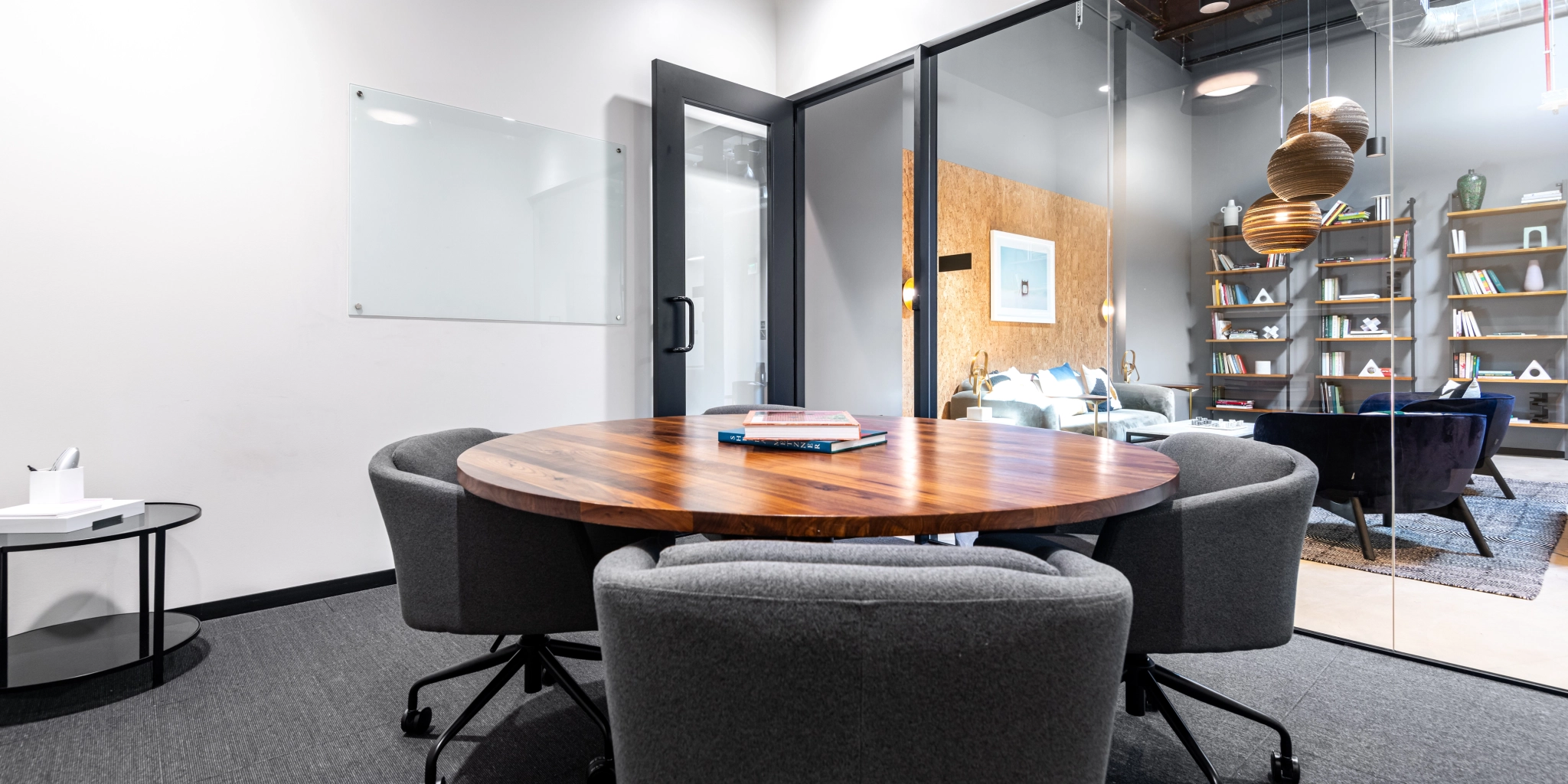 A modern coworking office space in Walnut Creek features a round wooden table, four gray chairs, a whiteboard on the wall, and an adjacent glass-walled room with shelves and pendant lights.
