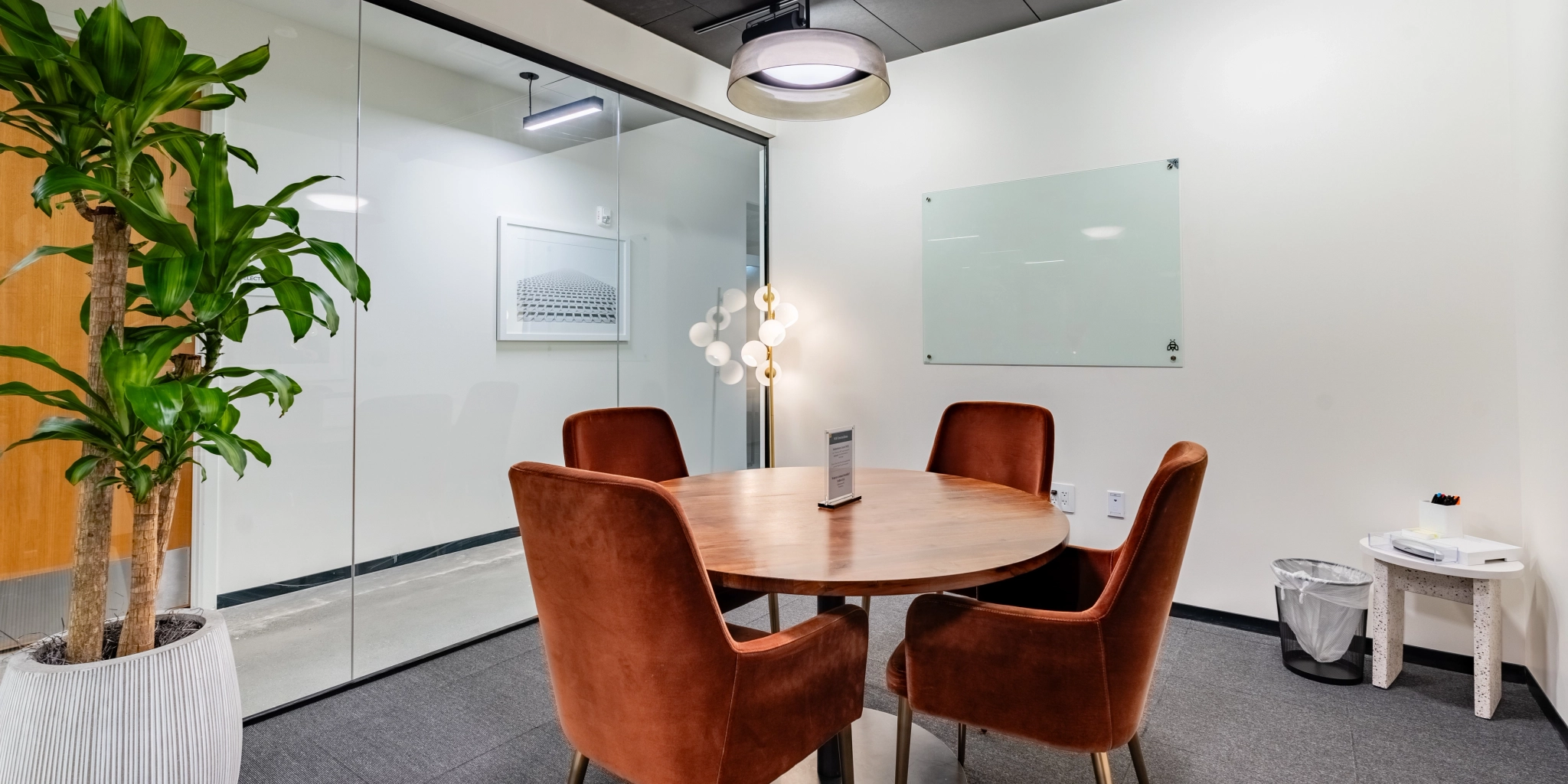 A modern Arlington office meeting room with a round wooden table, four brown chairs, a whiteboard, and a large plant in the corner. The workspace features glass walls and a minimalist design.