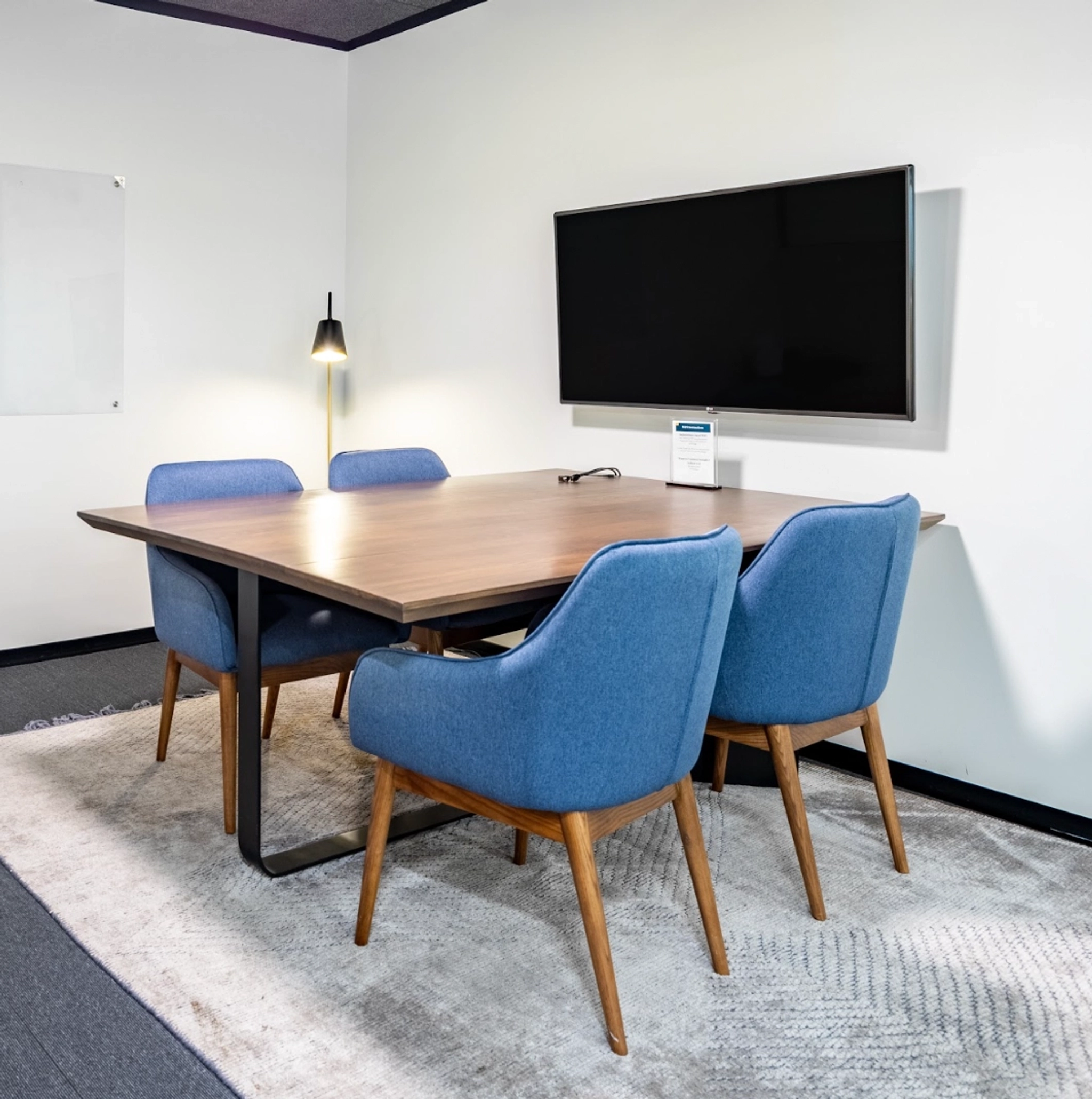 A small conference room, perfect for a productive workspace, features a rectangular wooden table, four blue upholstered chairs, a wall-mounted TV, a whiteboard, and a standing lamp.