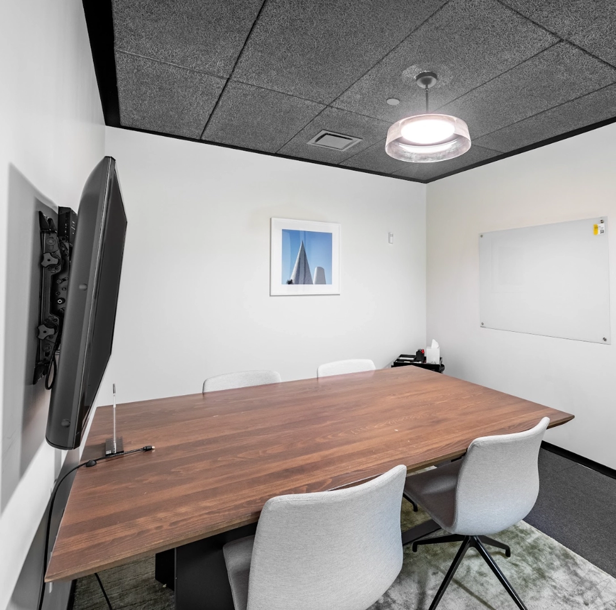 A small, modern coworking meeting room with a wooden table, four chairs, wall-mounted TV, framed wall art, ceiling light, and whiteboard.