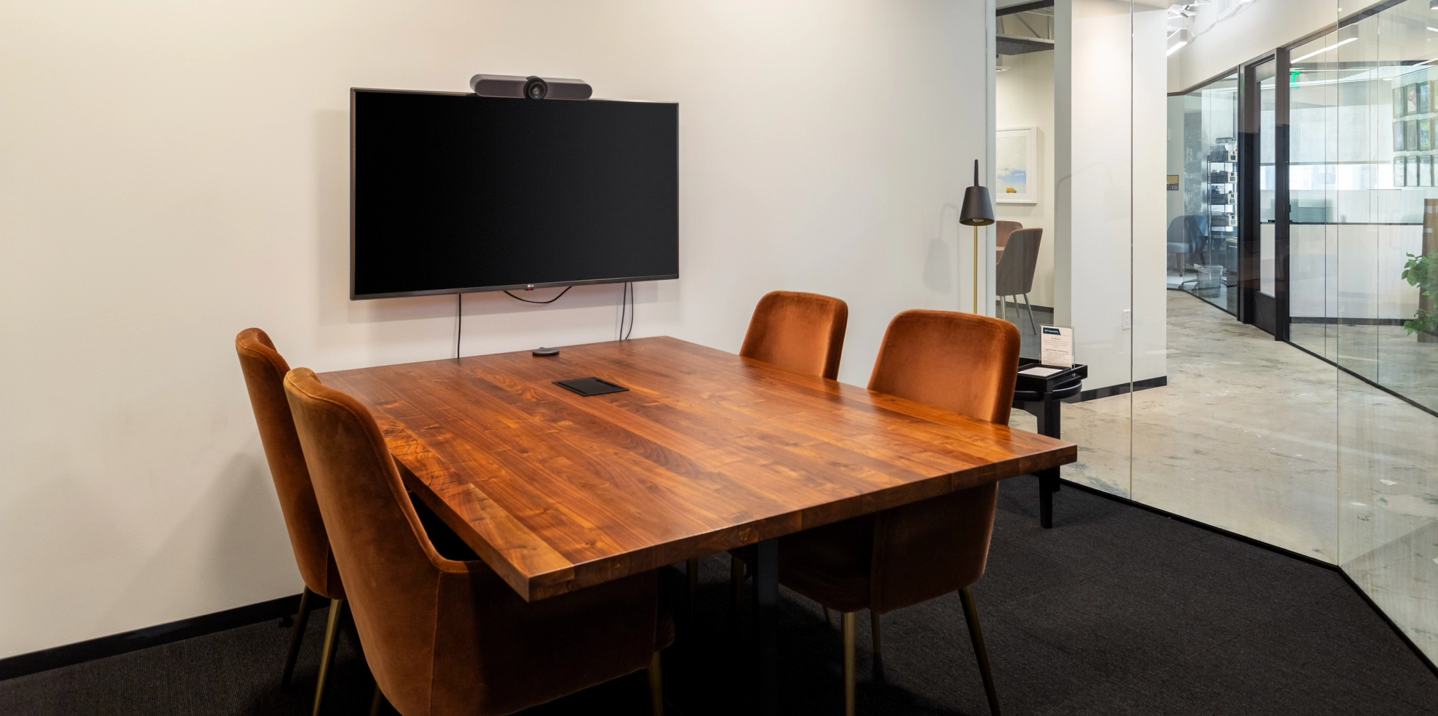 Een kleine, moderne vergaderruimte in San Francisco heeft een houten tafel, vier oranje stoelen, een aan de muur gemonteerd tv-scherm en een glazen wand.