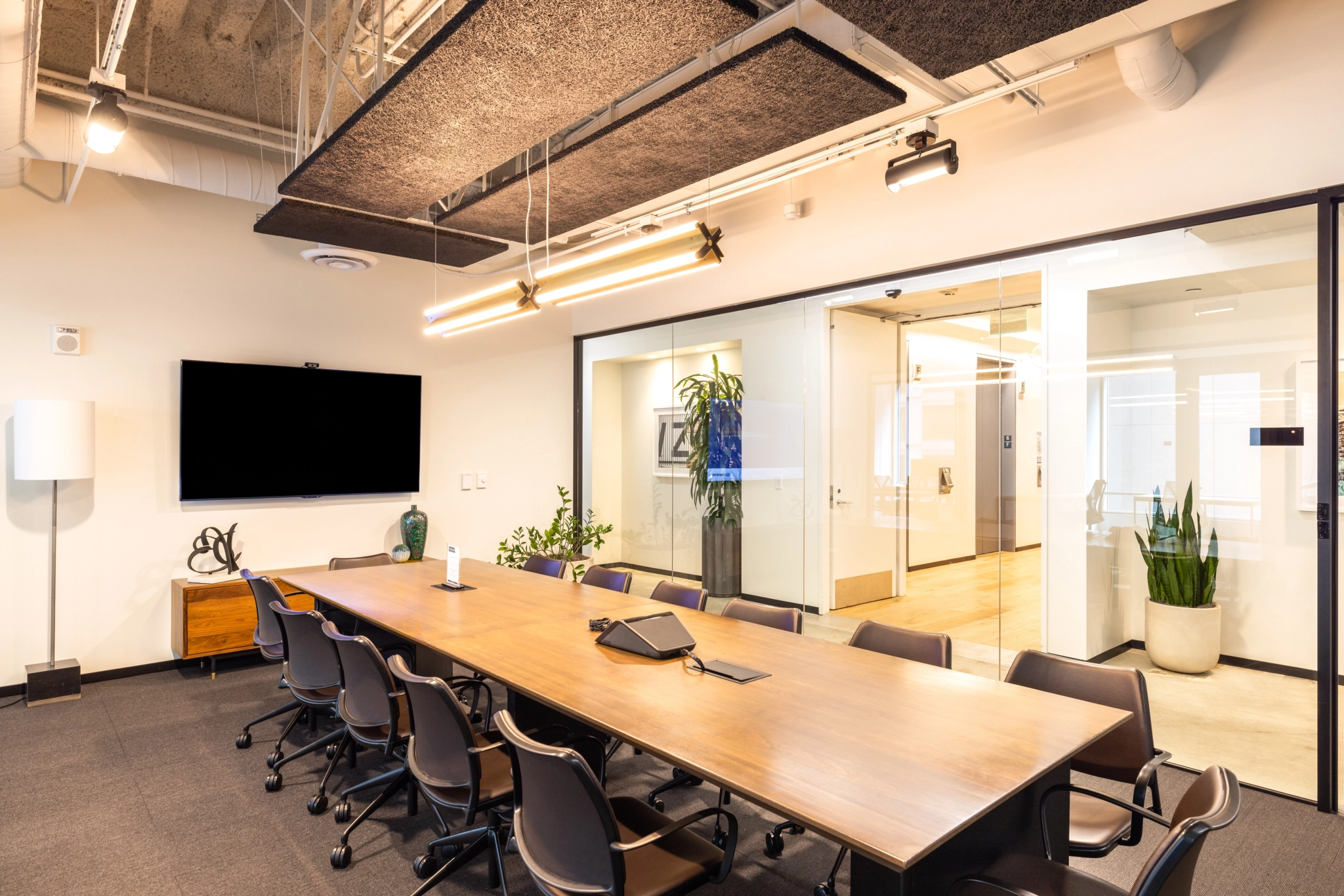 A modern coworking meeting room in San Francisco features a long wooden table, black chairs, a wall-mounted TV, and glass doors. Ceiling panels and sleek lighting fixtures are visible above, with a plant and cabinet nestled in the corner.