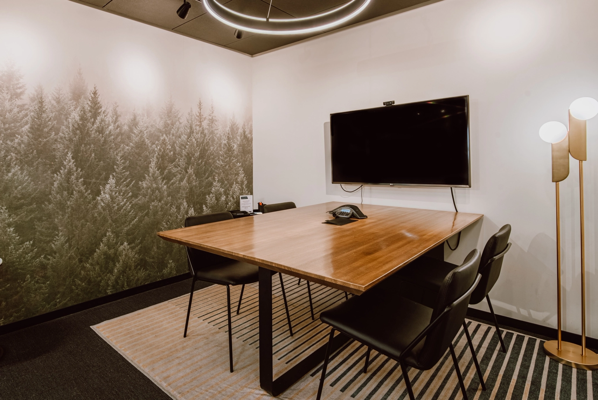 A modern meeting room with a wooden table, black chairs, and a large screen on the wall illuminated by a sleek lamp. The wall showcases a forest mural, creating an inspiring workspace.