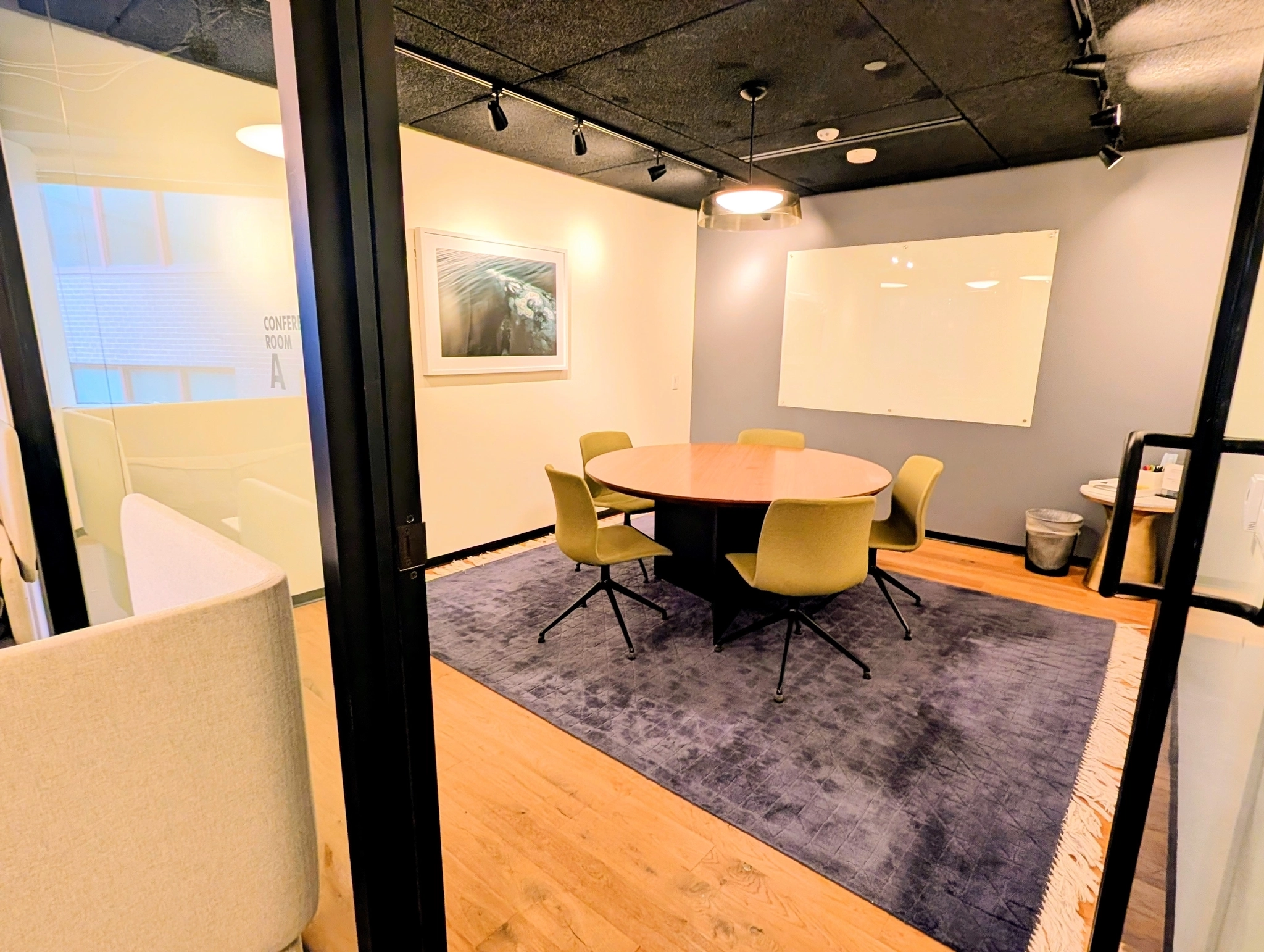 A modern Washington workspace conference room with a round table, five chairs, a whiteboard, wall art, and a blue rug on a wooden floor.