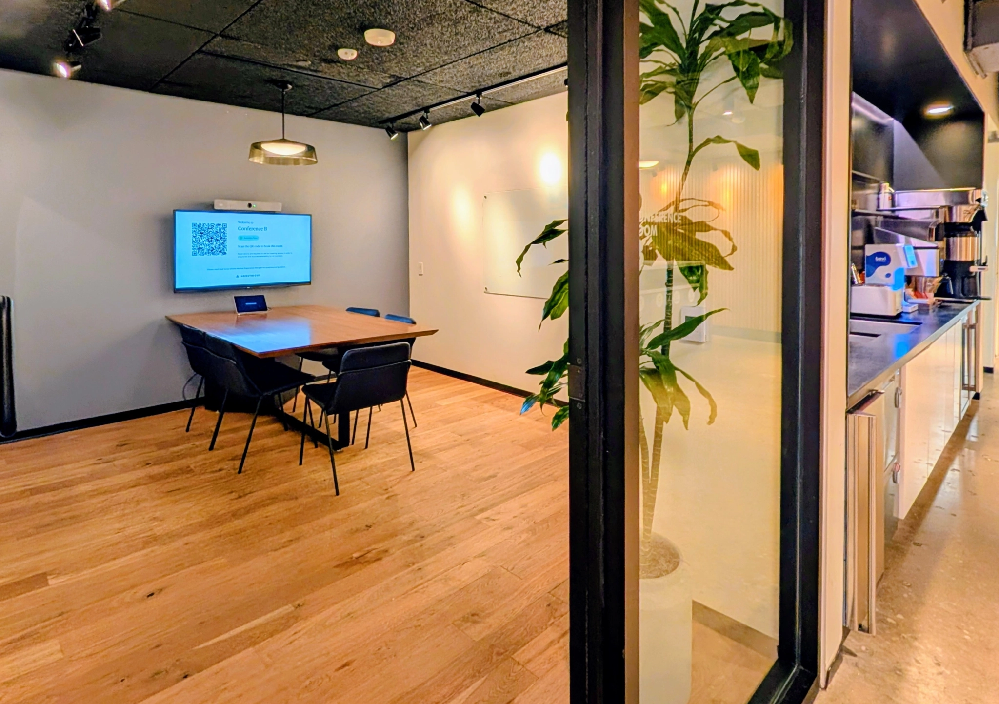 A modern coworking meeting room with a wooden table, five chairs, a wall-mounted screen displaying a QR code, a whiteboard, and a partial view of a kitchenette with a coffee machine.