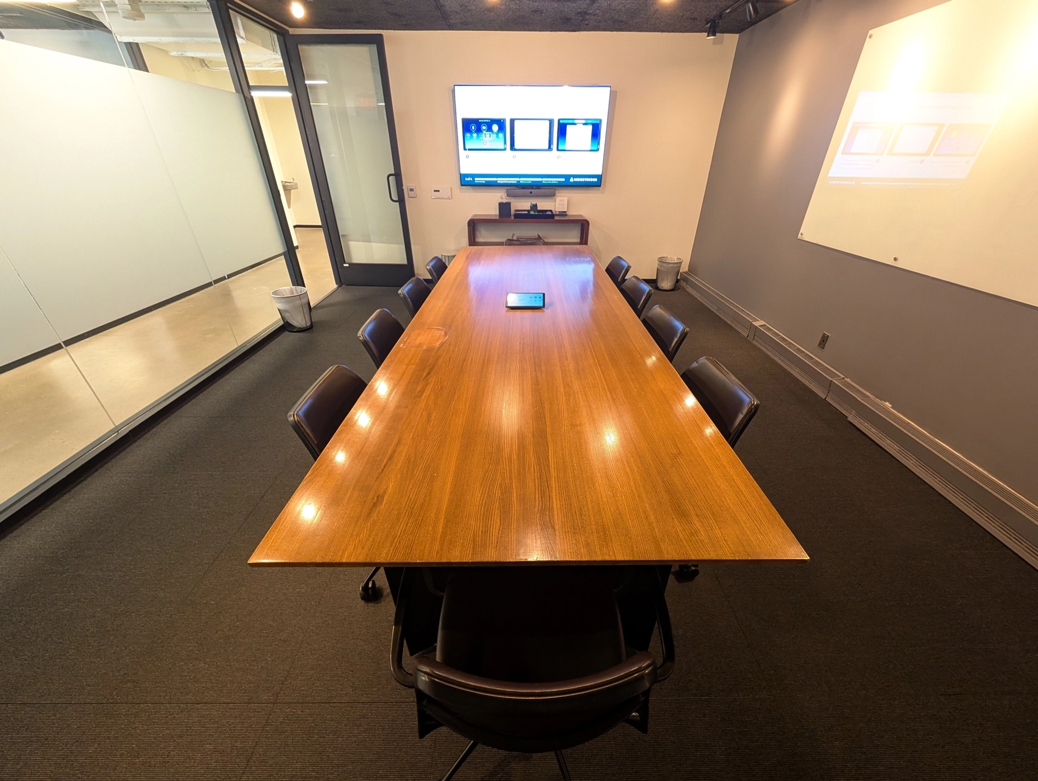 Een moderne vergaderruimte met een lange houten tafel, acht stoelen, een tv aan de muur, glazen wanden en een whiteboard, perfect voor coworking of kantoorvergaderingen.