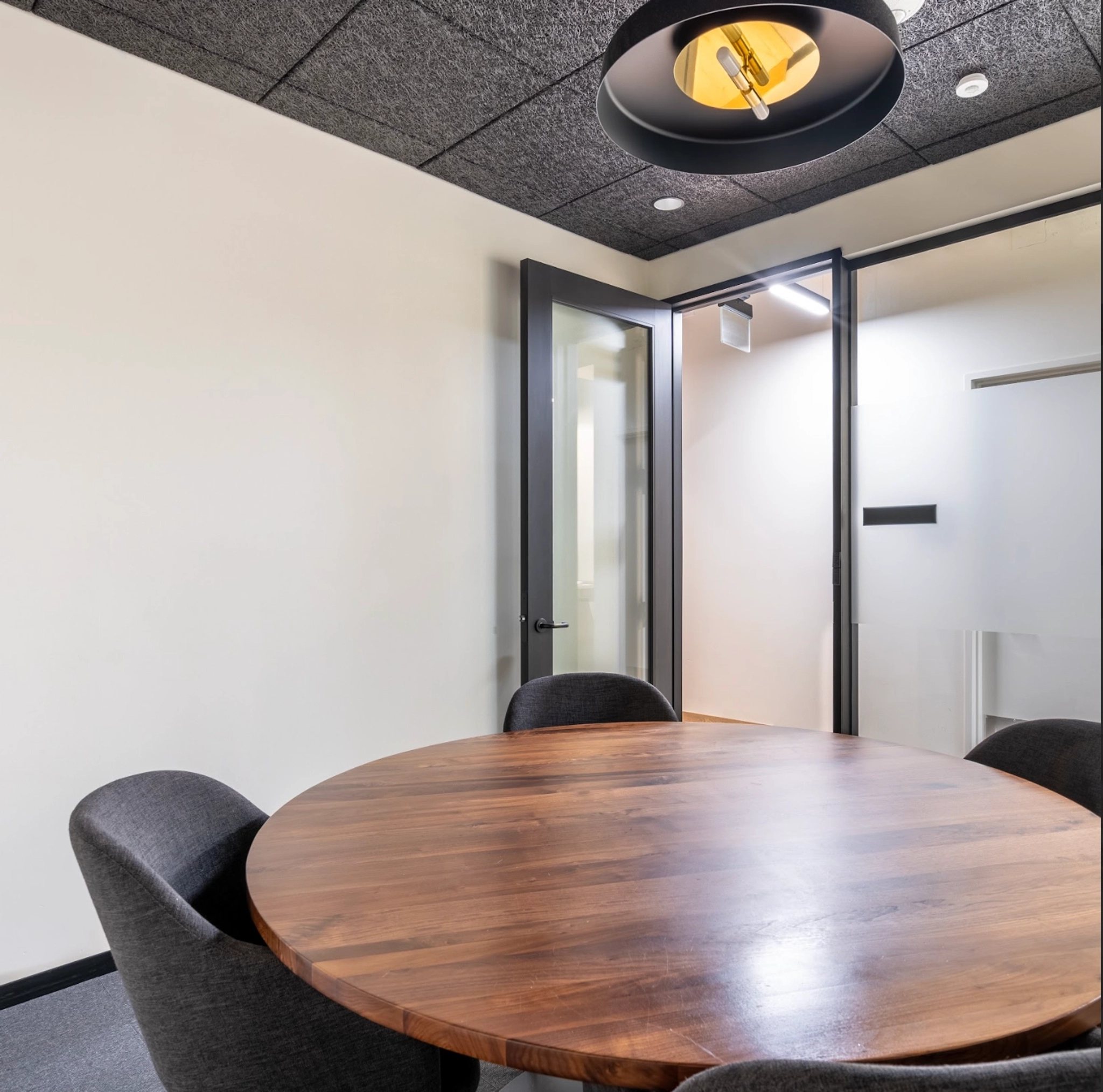 A small meeting room in the Concord office features a round wooden table surrounded by four dark chairs, a glass door slightly open, and a modern ceiling light fixture above.