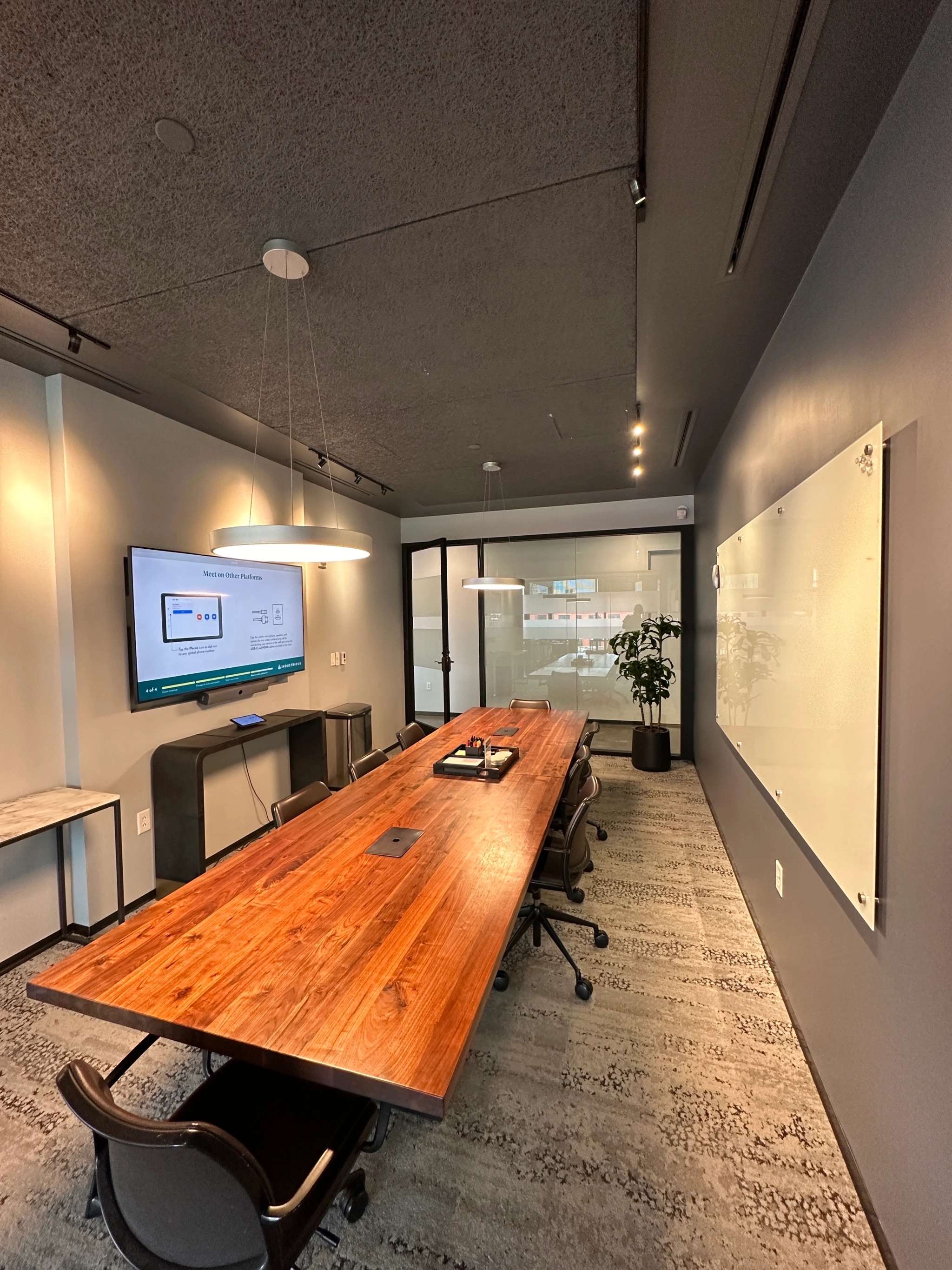 A modern meeting room featuring a long wooden table with chairs, a wall-mounted TV displaying a presentation, and a whiteboard. A plant is positioned near the glass door at the far end of this coworking space.