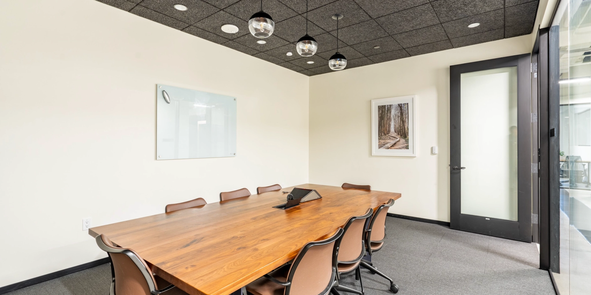 Een moderne coworking-conferentieruimte met een houten tafel, acht stoelen, een glazen deur, een whiteboard en een ingelijste foto aan de muur. Aan het plafond zijn hanglampen zichtbaar.