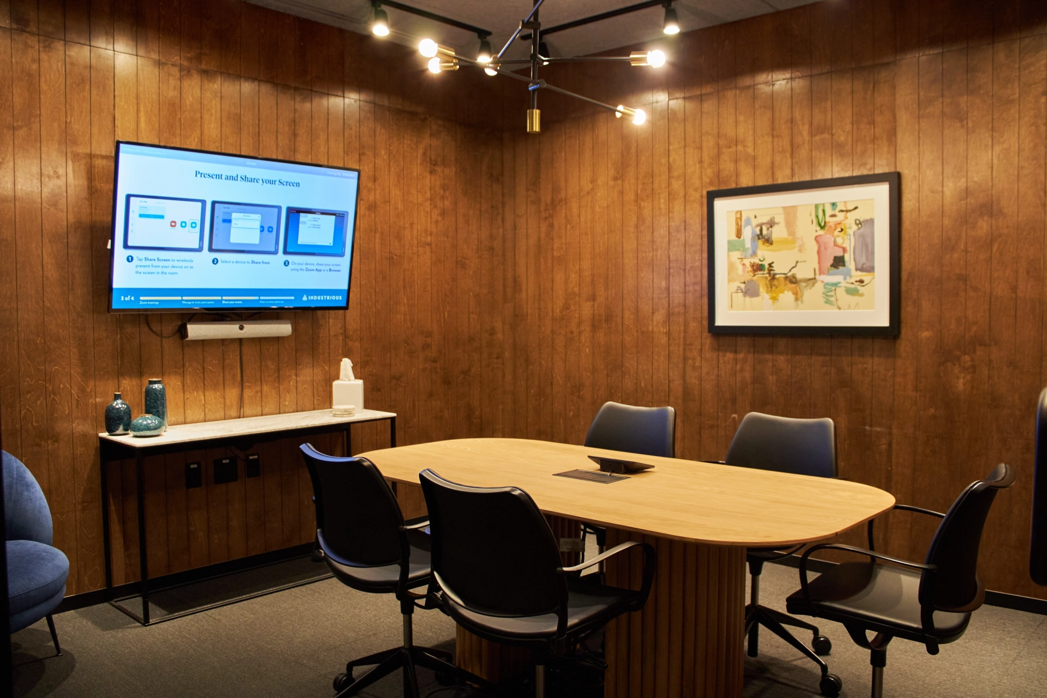 A meeting room with a wooden table, chairs, wall-mounted TV screen displaying a presentation, and framed artwork on a wooden wall.