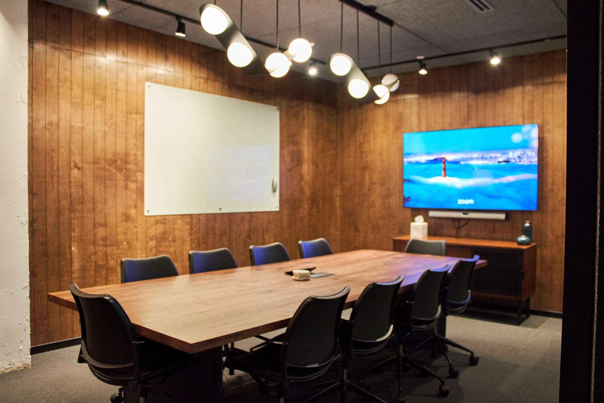 Modern conference room in a coworking space with a wooden table, eight chairs, a wall-mounted whiteboard, a large screen displaying the Zoom interface, and a unique overhead light fixture.