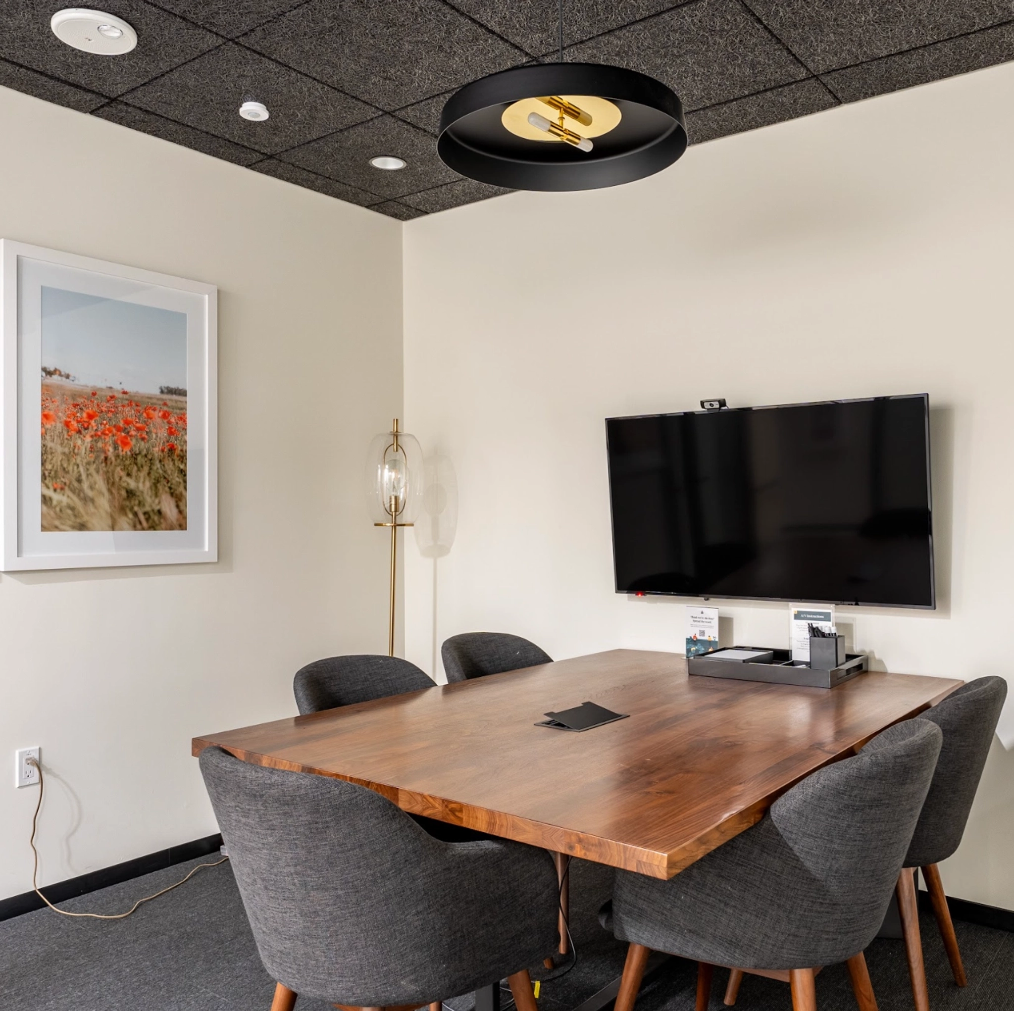 A small meeting room with a wooden table, four gray chairs, a wall-mounted TV, and a framed picture. This cozy workspace also features a unique light fixture on the ceiling and a floor lamp.