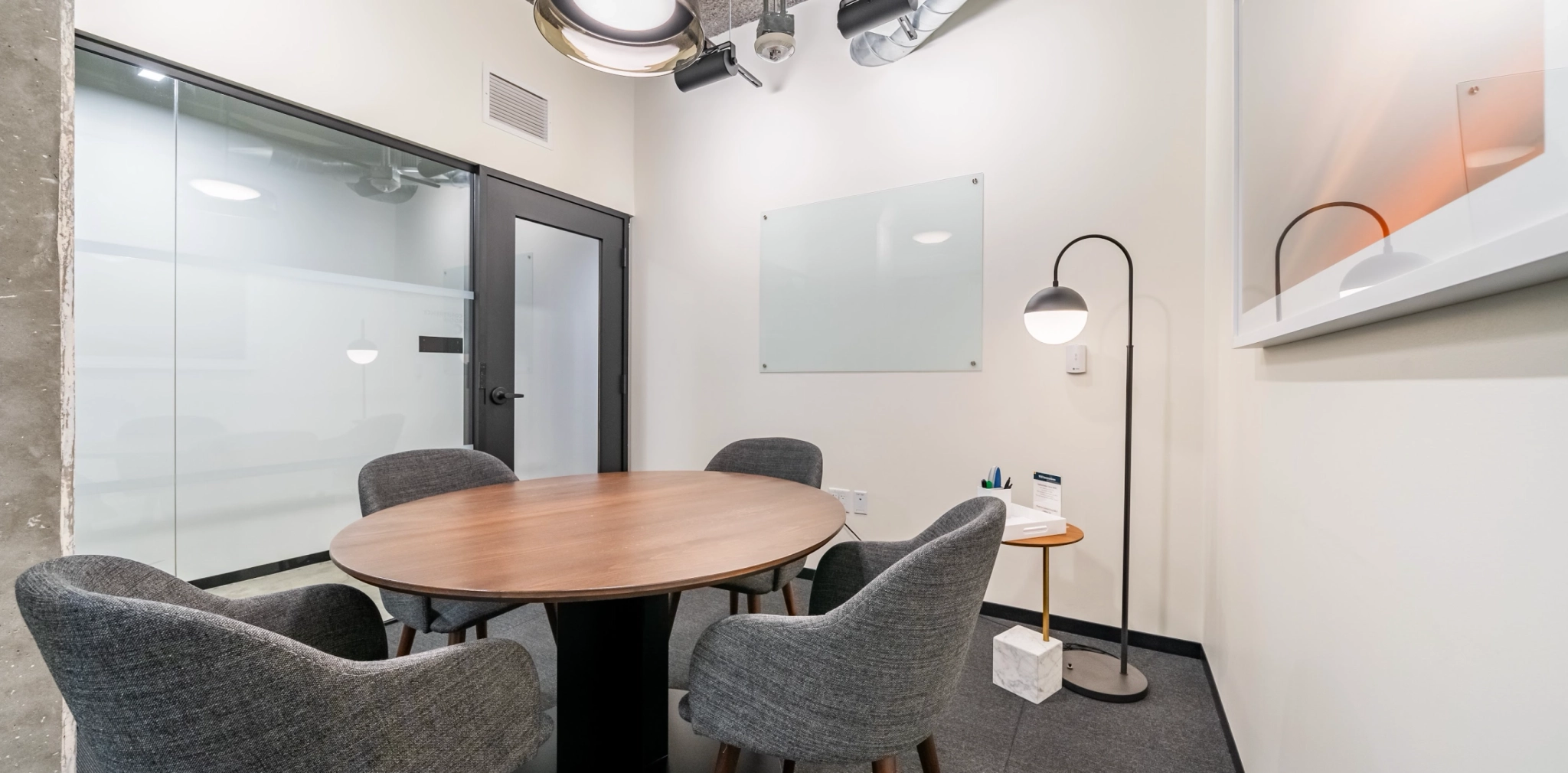 This cozy office meeting room features a round wooden table surrounded by four gray chairs, perfect for collaborative discussions. A whiteboard adorns the wall, complemented by a floor lamp. To the left, a door with frosted glass leads to the rest of the coworking space.