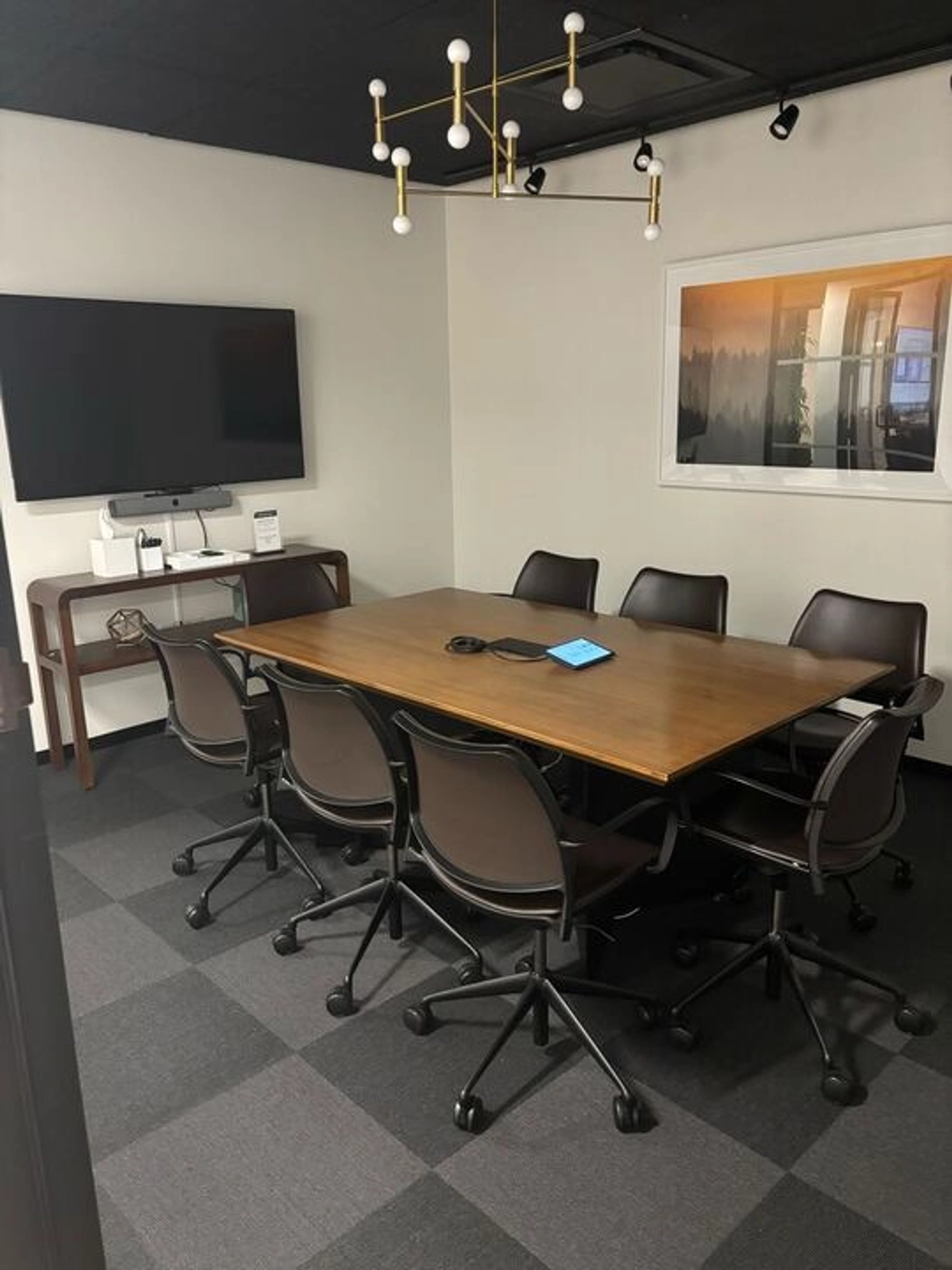 A meeting room in Boston features a wooden table surrounded by six chairs. A large TV is mounted on the wall, accompanied by a ceiling light fixture and a framed picture, creating an ideal workspace for collaboration.