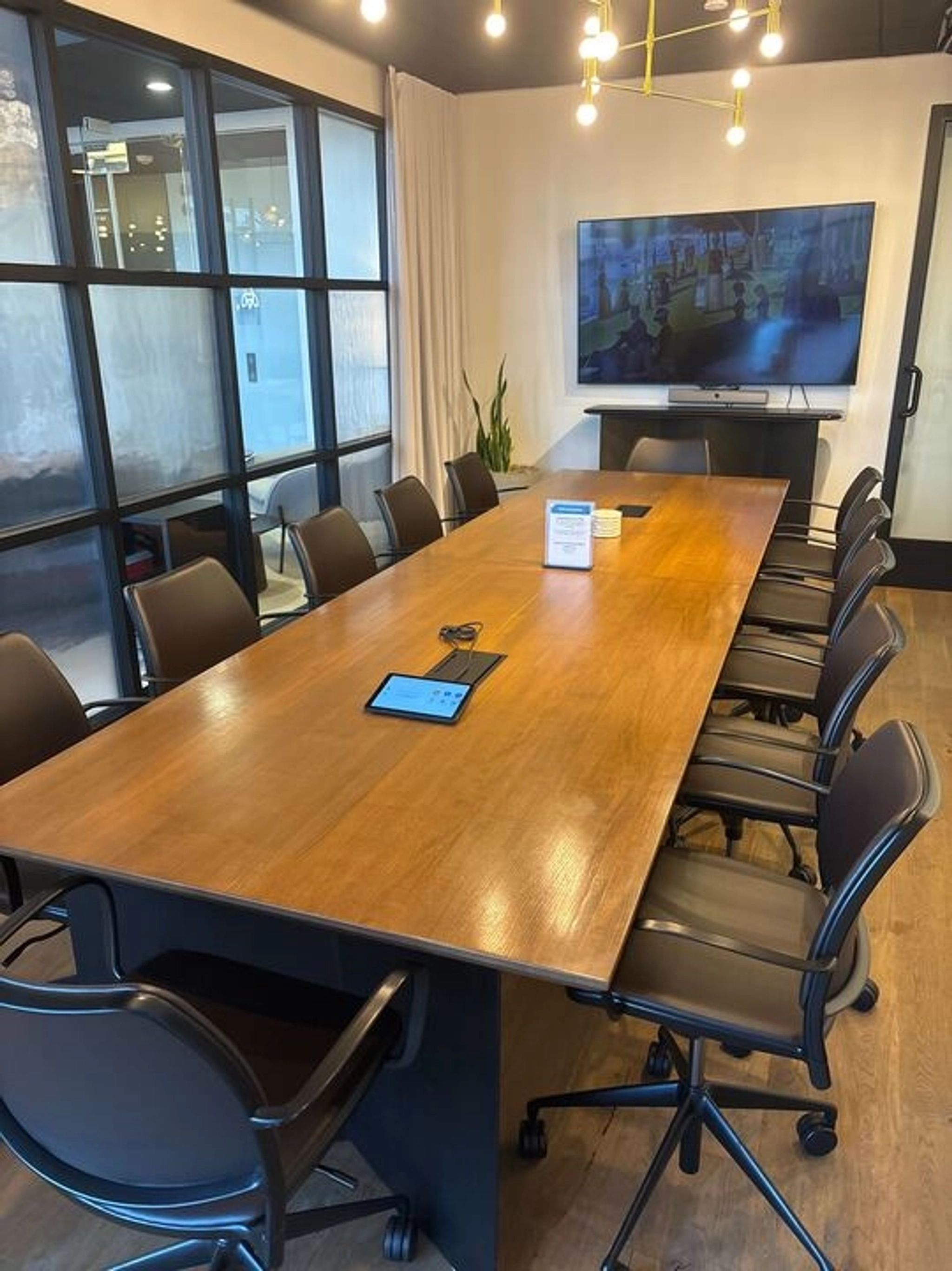 A modern meeting room with a long wooden table, ten chairs, a wall-mounted screen, and a hanging light fixture. Perfect for coworking in the heart of Boston, it features a tablet and documents ready on the table for your next collaboration.