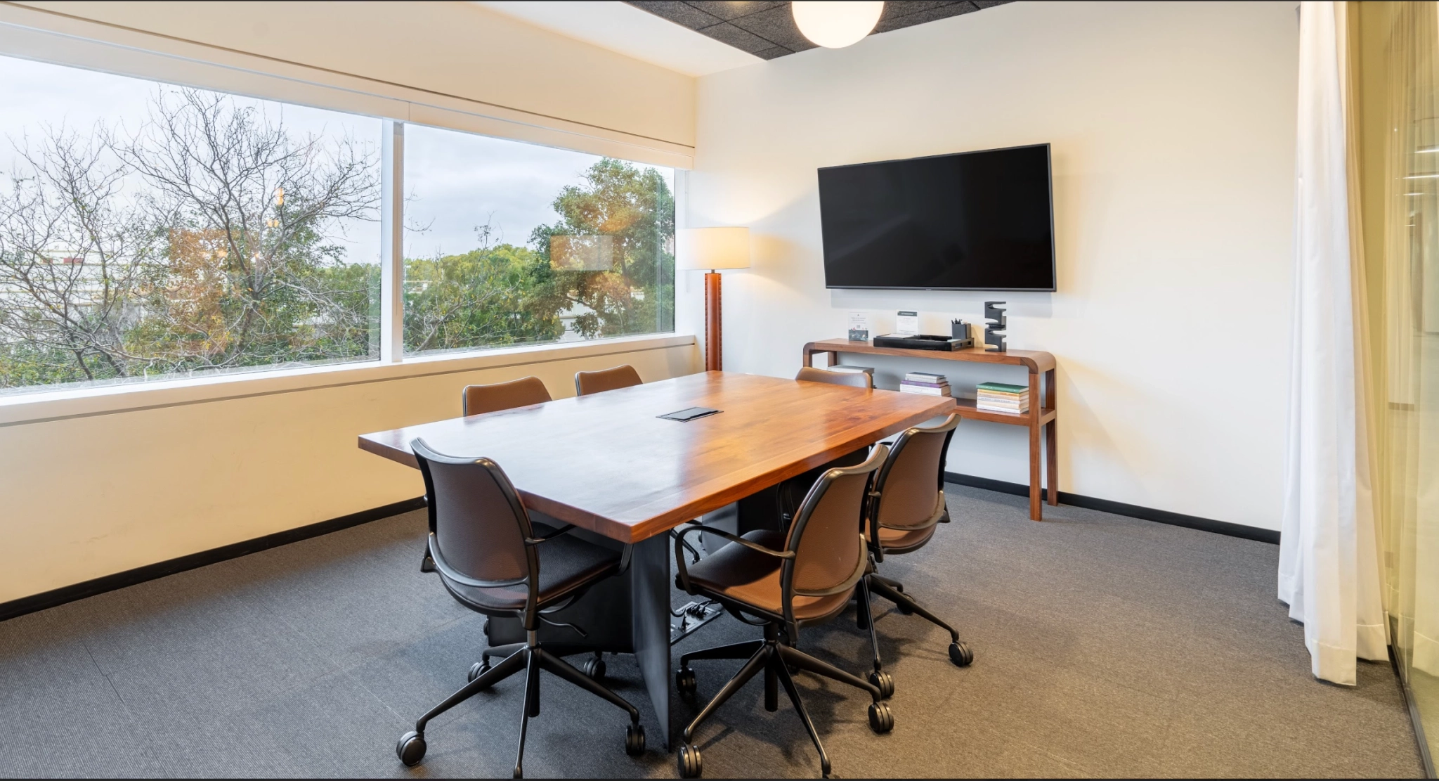 Une petite salle de conférence avec une table en bois entourée de six chaises de bureau, une télévision murale et une grande fenêtre donnant sur les arbres. Cet espace de travail de Concord comprend également une table d'appoint avec des livres et du matériel contre le mur, parfaite pour les séances de coworking.