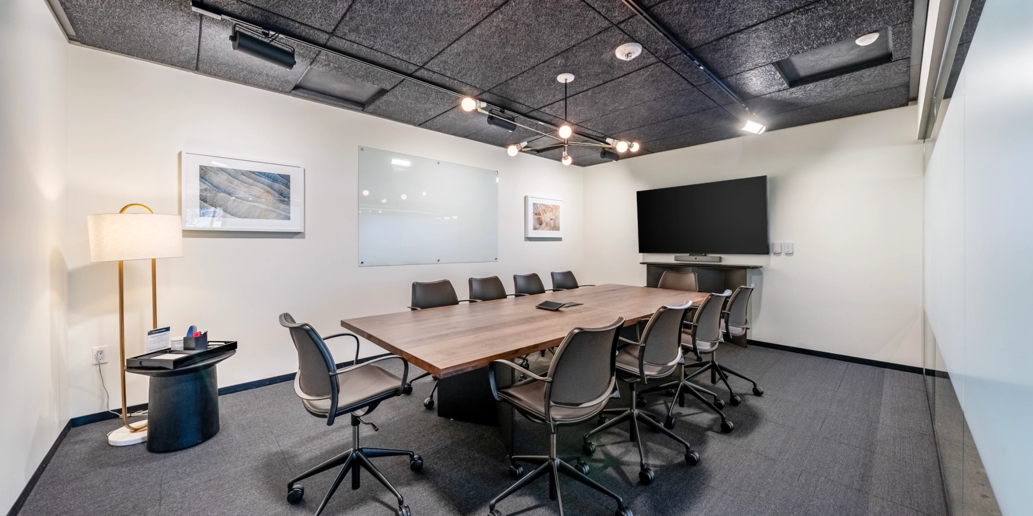 A modern meeting room in San Francisco, featuring a long wooden table, ten chairs, a large screen, a whiteboard, framed artwork, and a floor lamp.