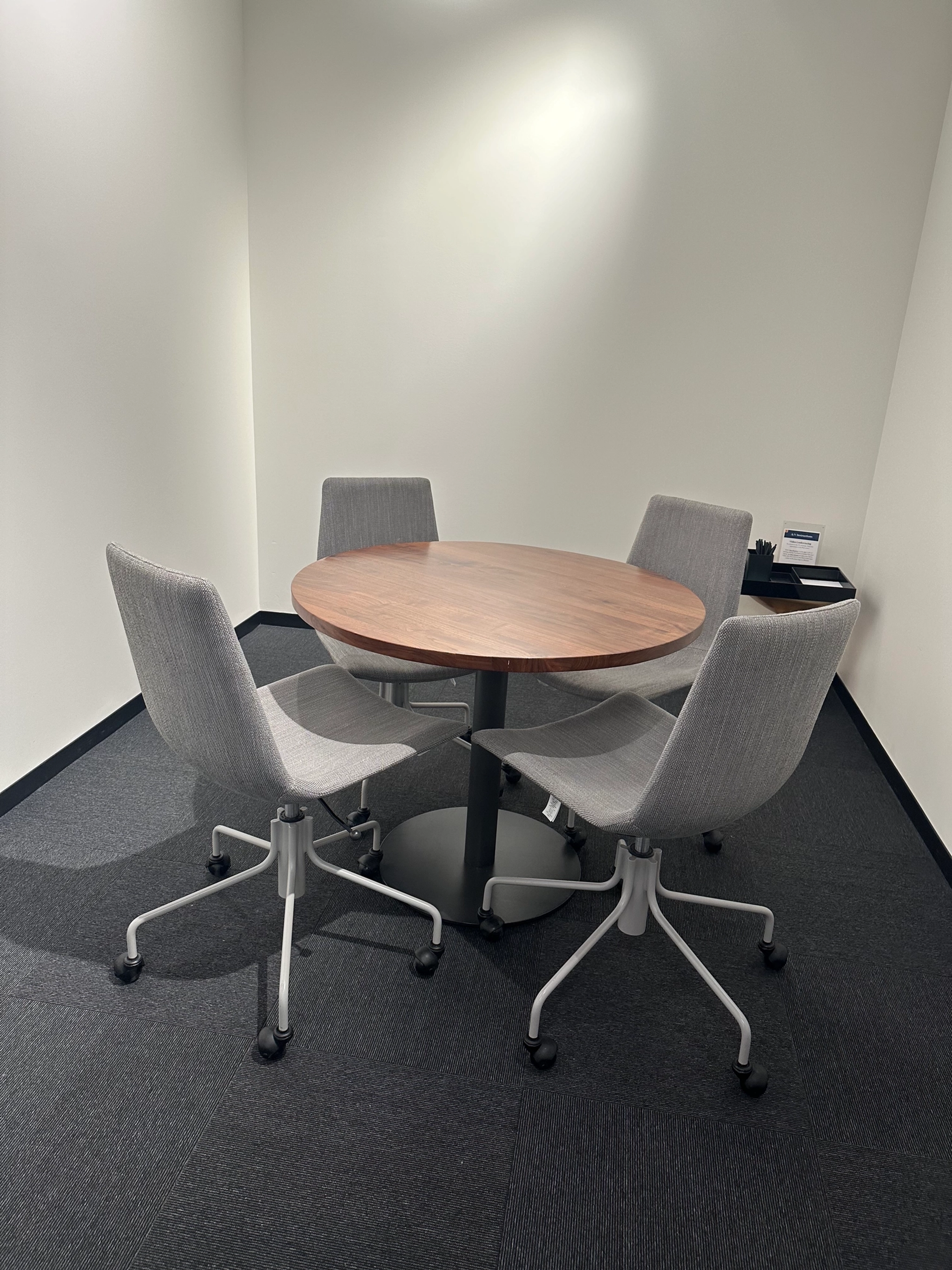 Un petit espace de travail à New York avec une table ronde en bois et quatre chaises de bureau grises sur un sol recouvert de moquette foncée. Un téléphone et un bloc-notes sont posés sur une petite table d'angle, parfaite pour des réunions productives.