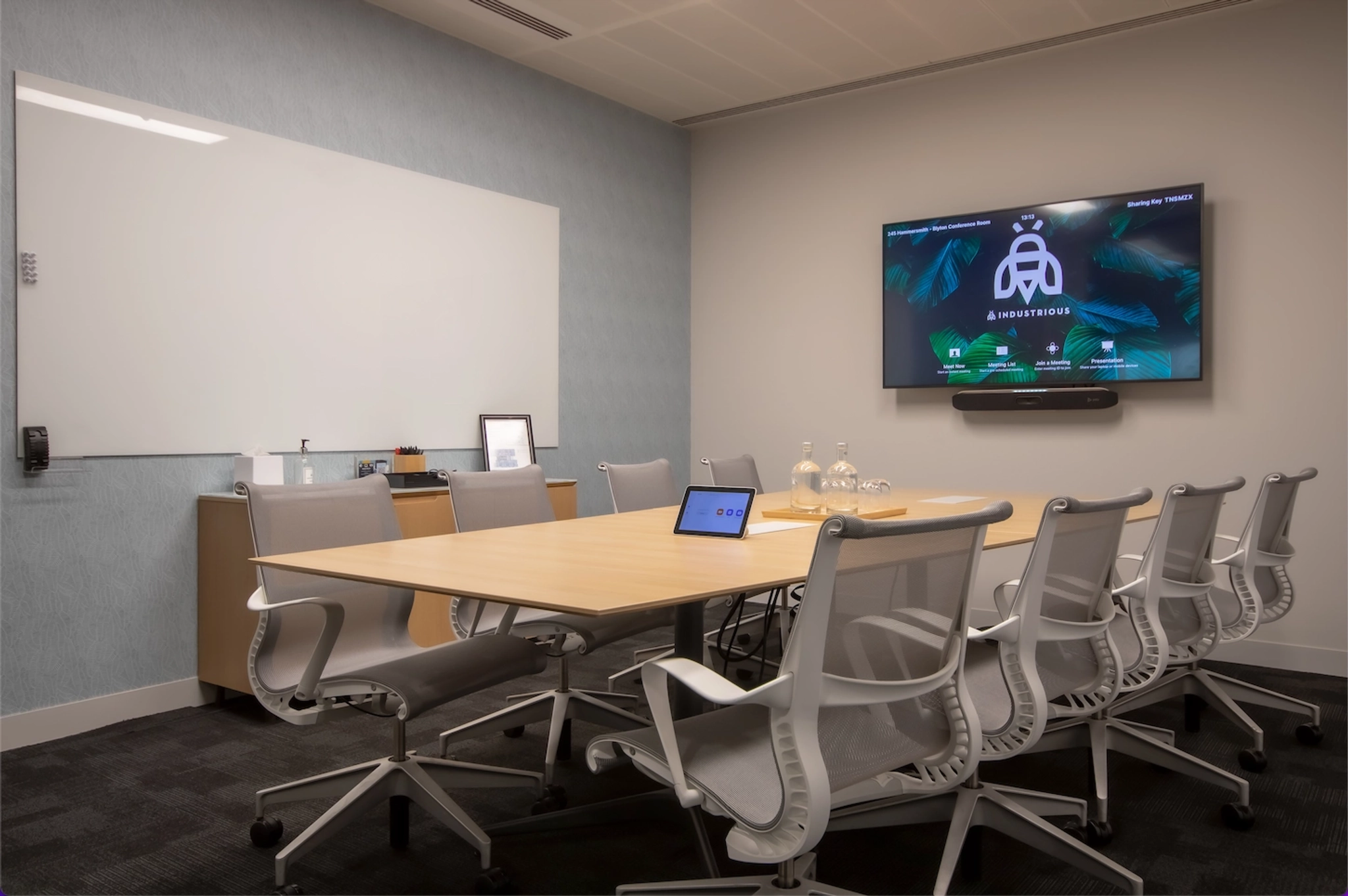 A modern meeting room with a long table, eight office chairs, a wall-mounted TV screen displaying a logo and interface, and a whiteboard. An electronic device is placed on the table, enhancing this coworking space for productivity.