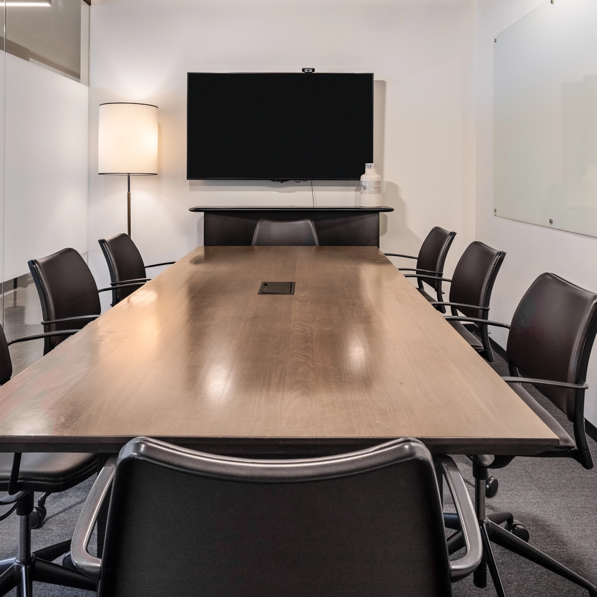 Une salle de conférence dans le bureau de coworking comprend une longue table en bois entourée de huit chaises, une télévision murale, une lampe dans le coin et un tableau blanc sur le mur.