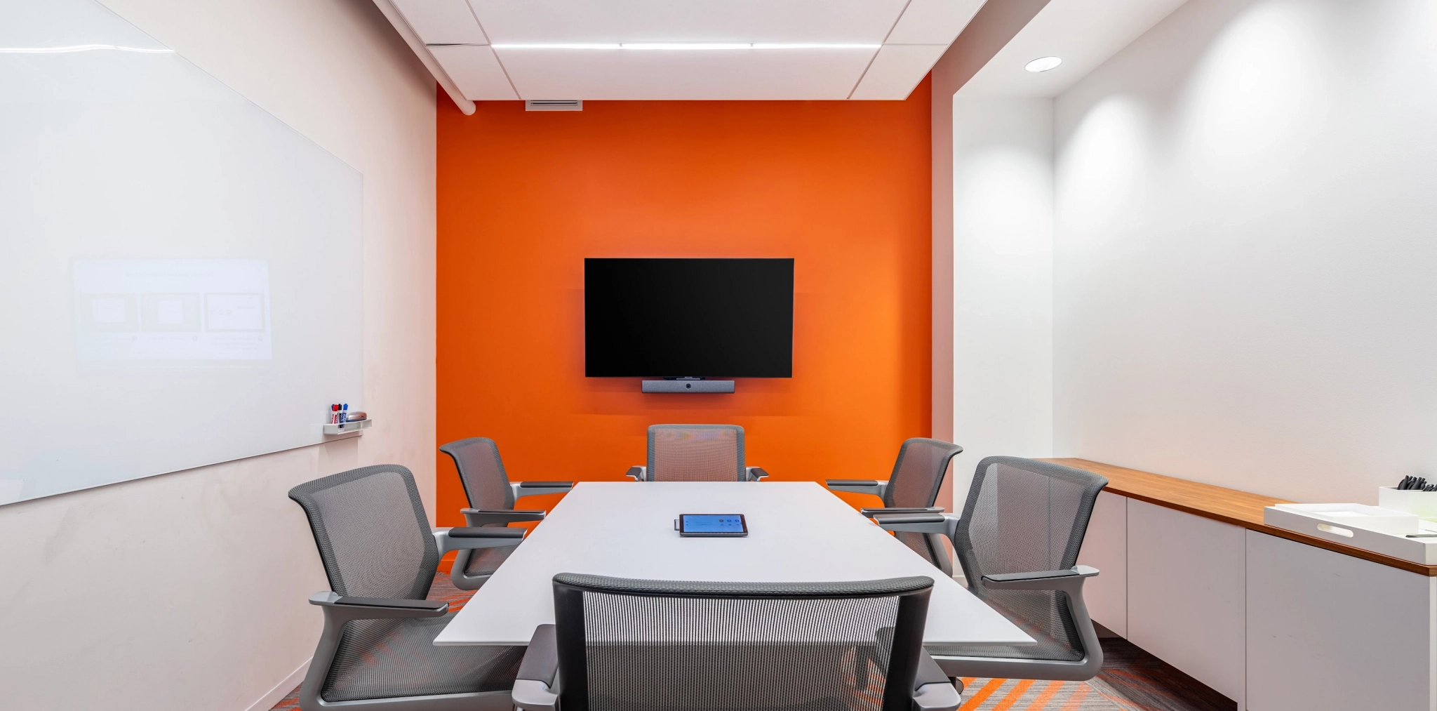 A modern coworking conference room with an orange accent wall, a whiteboard on the left, a wall-mounted TV on the far wall, and a rectangular table surrounded by six office chairs. A tablet is placed on the table.