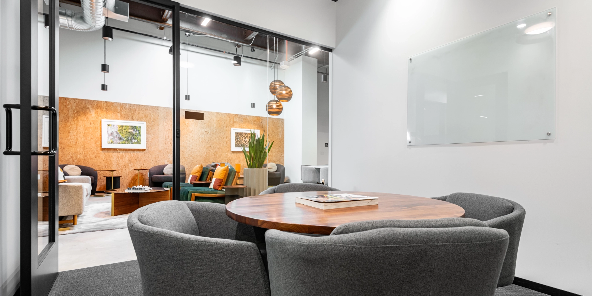 A small coworking room with a round wooden table and four gray chairs, adjacent to a larger lounge area. The workspace features a glass wall with a door and a whiteboard on the wall.