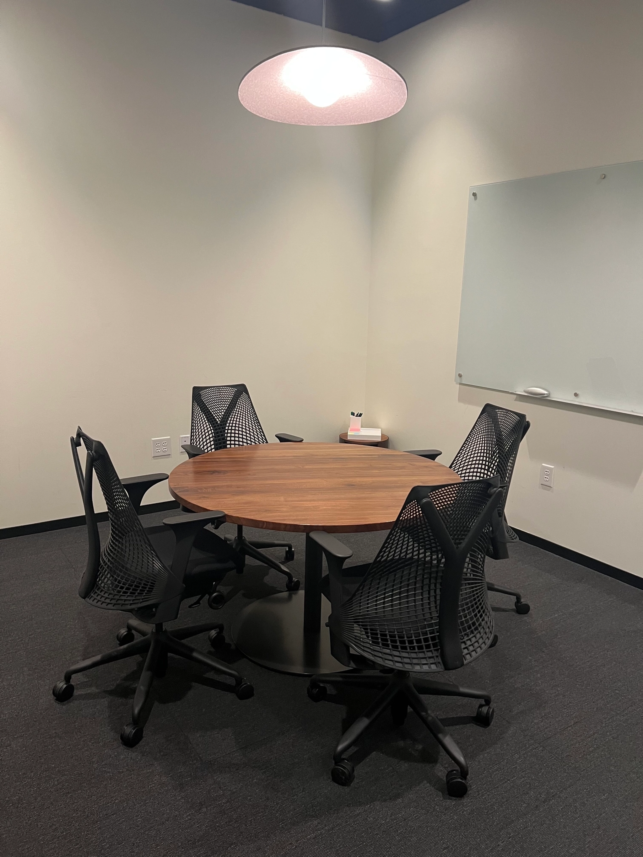 Une petite salle de réunion avec une table ronde en bois, quatre chaises de bureau, un tableau blanc au mur et un plafonnier. L'espace de travail présente une palette de couleurs neutres et de la moquette.