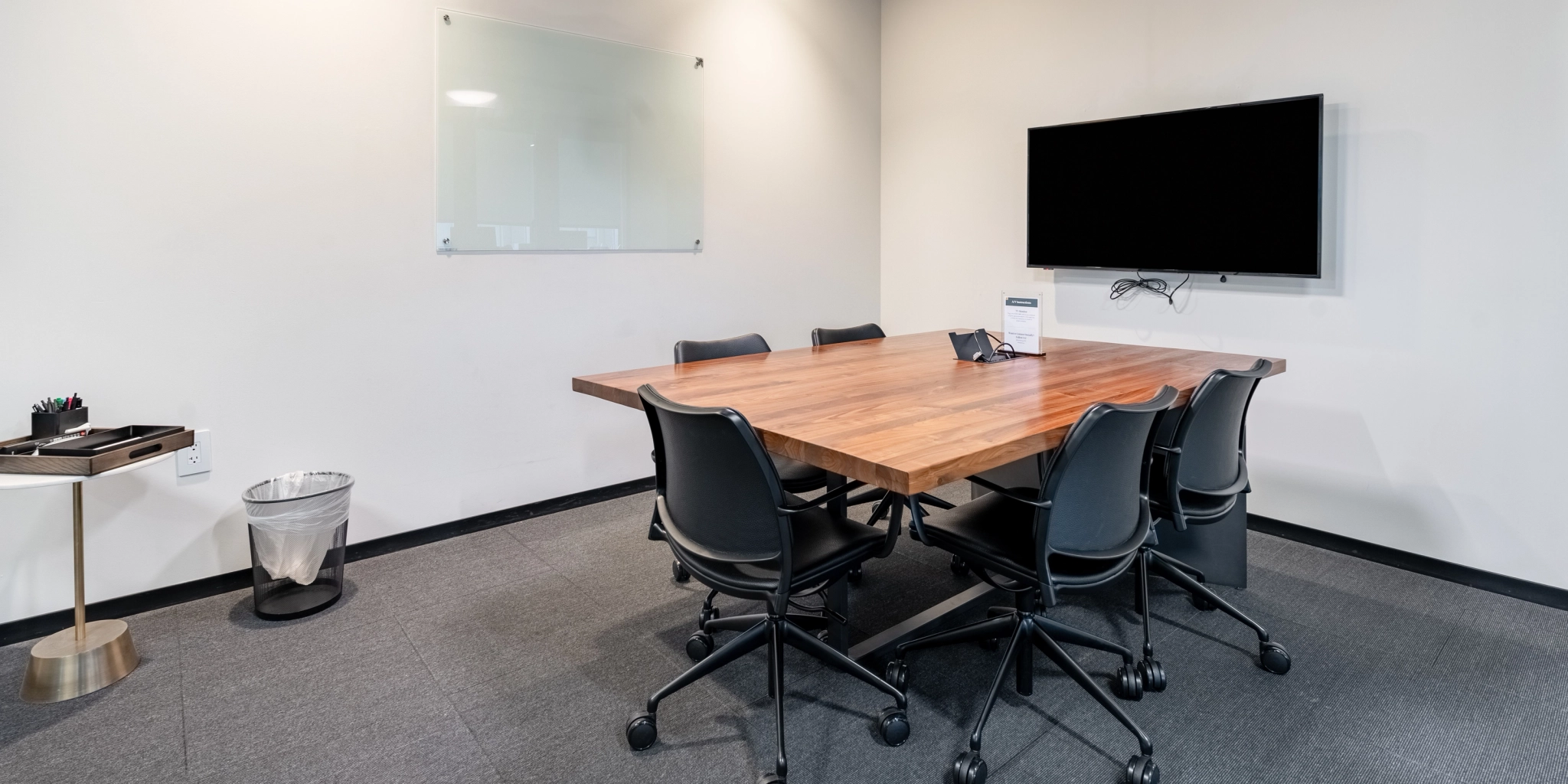 Een moderne vergaderruimte in de werkruimte van Arlington is voorzien van een houten tafel, zes stoelen, een aan de muur gemonteerde tv, een glazen whiteboard, een vuilnisbak en een klein bijzettafeltje met kantoorbenodigdheden.