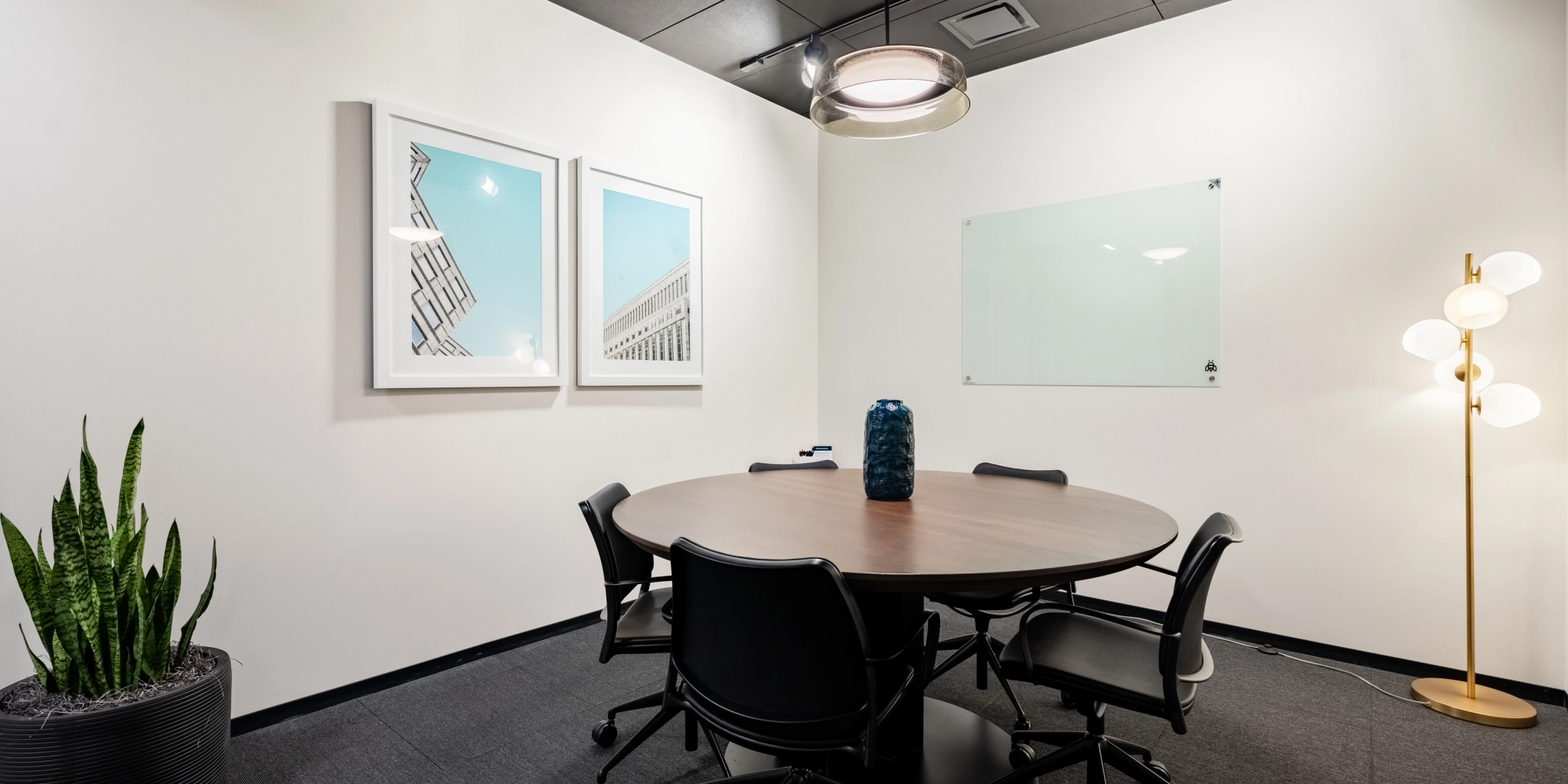 A small conference room in the Arlington office, featuring a round table surrounded by four chairs, a potted plant, framed wall art, a glass board for brainstorming sessions, a floor lamp, and a ceiling light fixture—perfect for creating an inspiring workspace.