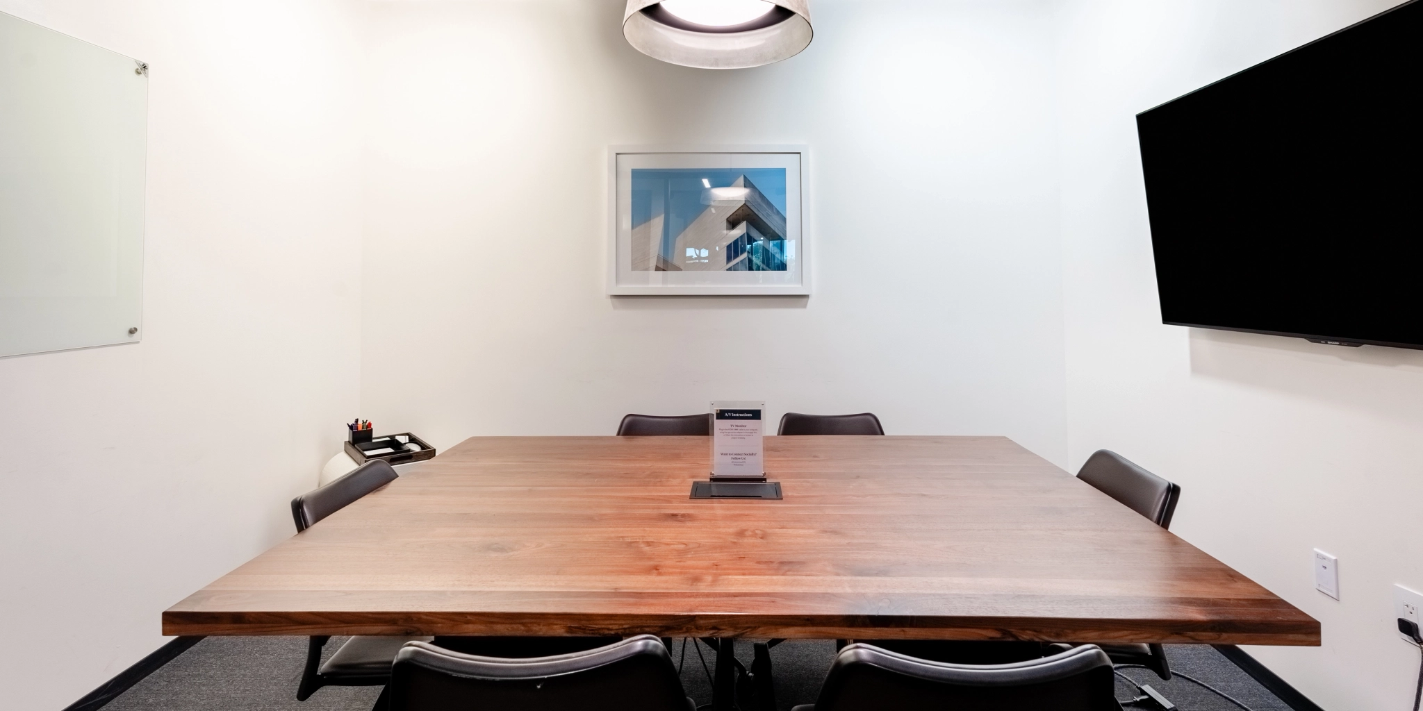 A coworking space featuring a wooden table, six chairs, a wall-mounted screen, a framed picture, a light fixture, and a phone on the side.