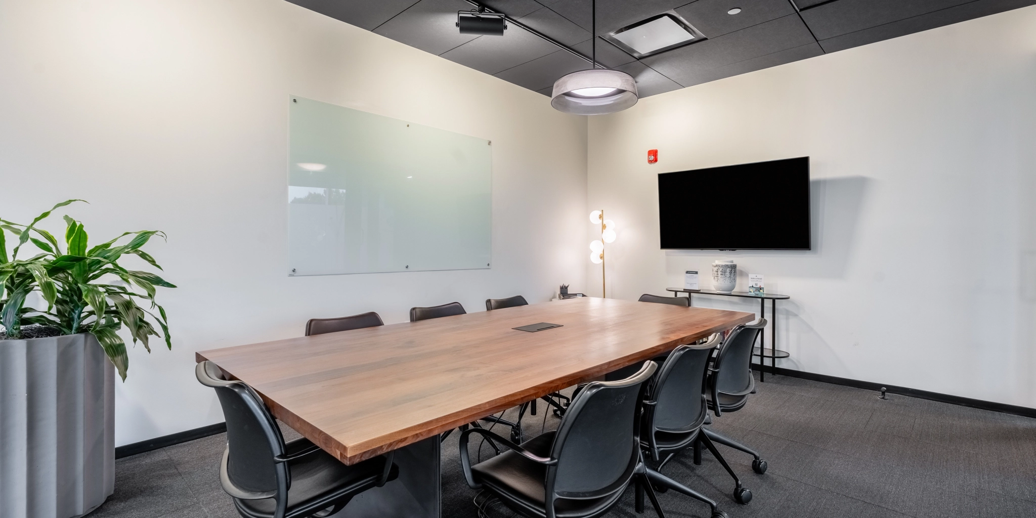 Une salle de conférence de bureau moderne à Arlington comprend une grande table en bois entourée de chaises, un tableau blanc vierge sur le mur, une télévision à écran plat et une plante dans le coin, créant un espace de travail idéal.