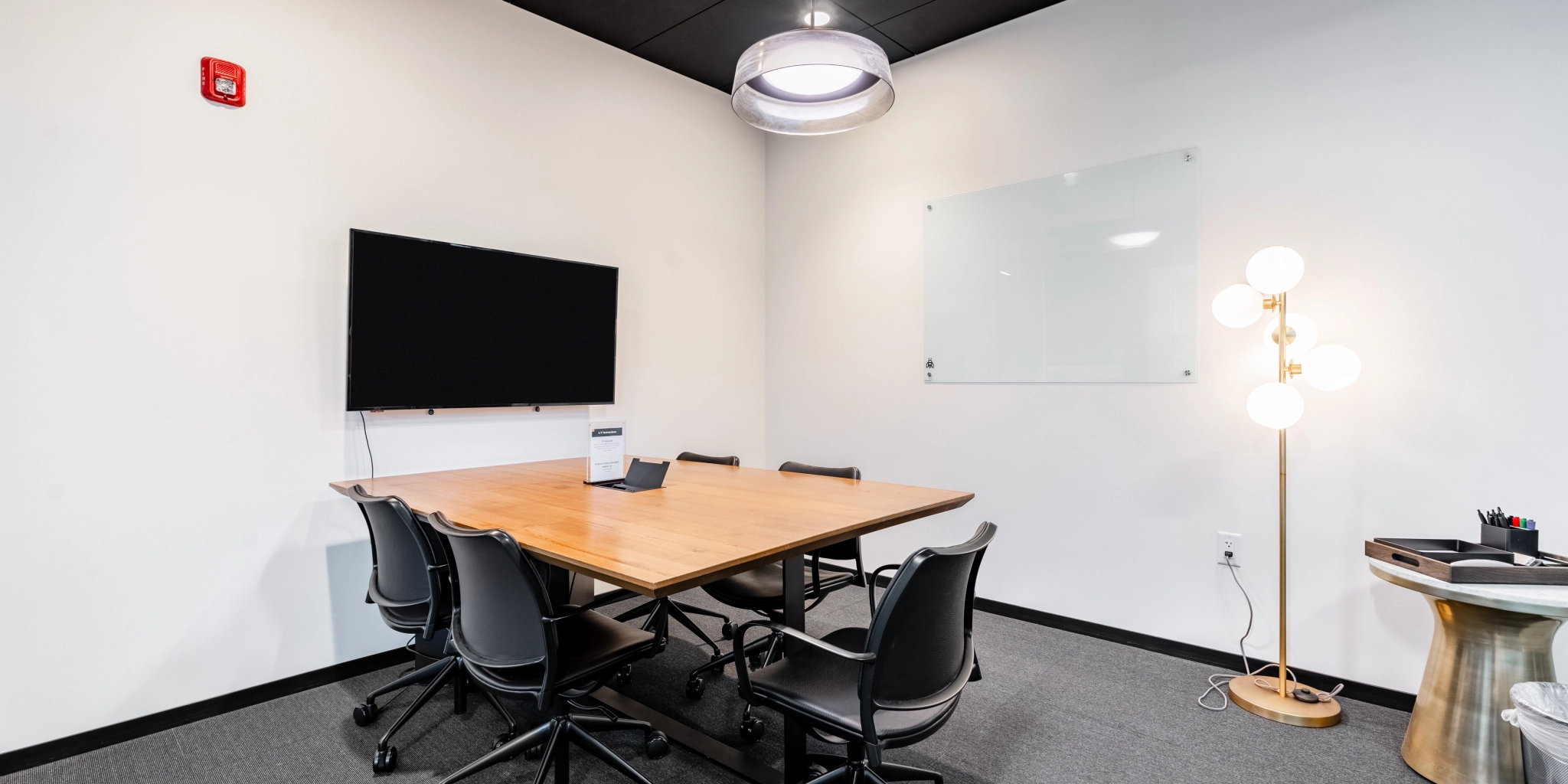 Een kleine vergaderruimte in Arlington heeft een houten tafel, zwarte stoelen, een aan de muur gemonteerde tv en een whiteboard. De lamp wordt verlicht door een vloerlamp met meerdere lampen en bevat essentiële kantoorbenodigdheden, netjes gerangschikt op een bijzettafel.